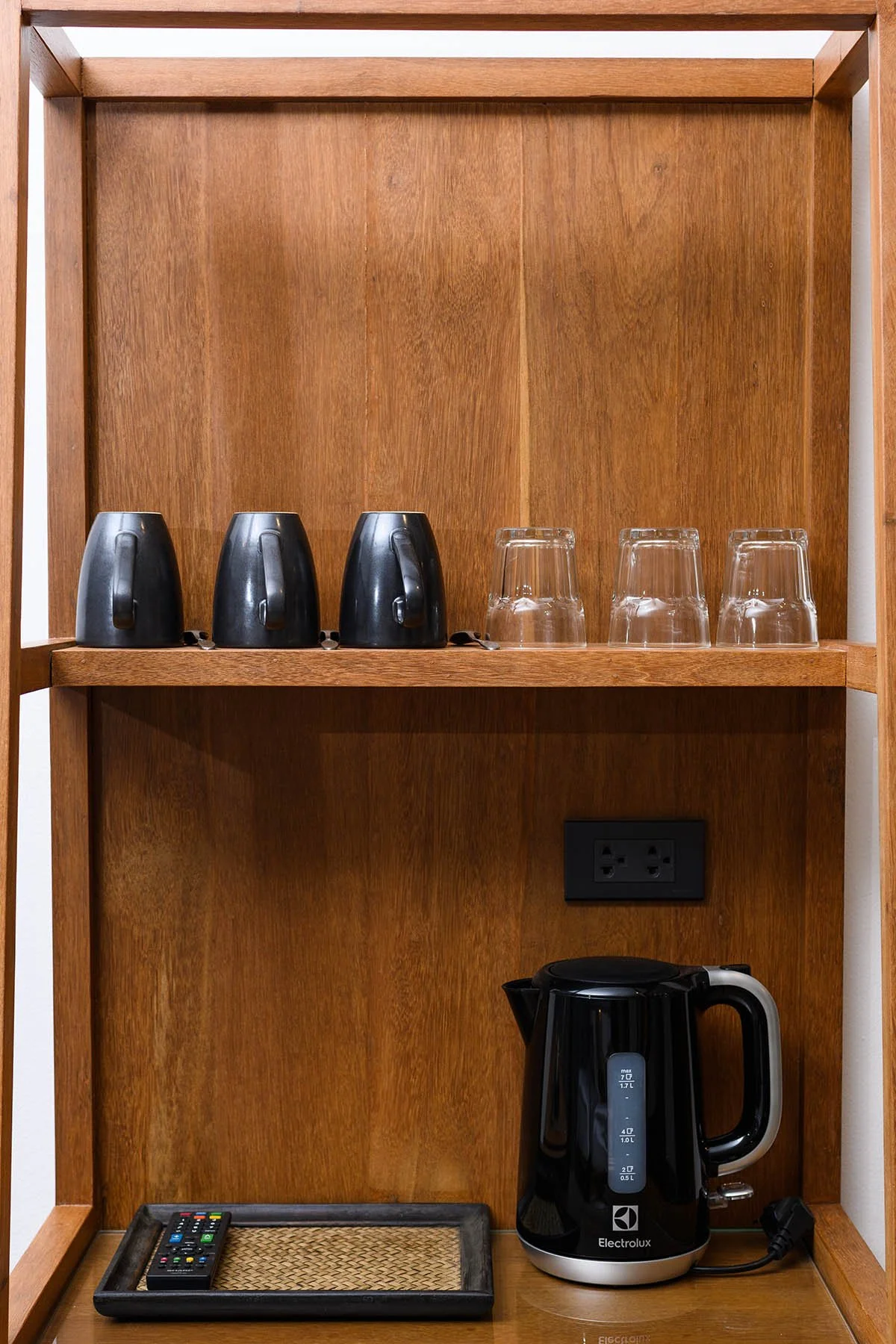 Wooden shelf with three black coffee mugs, three clear glasses, a black electric kettle, a tray with a remote control, and a power outlet.