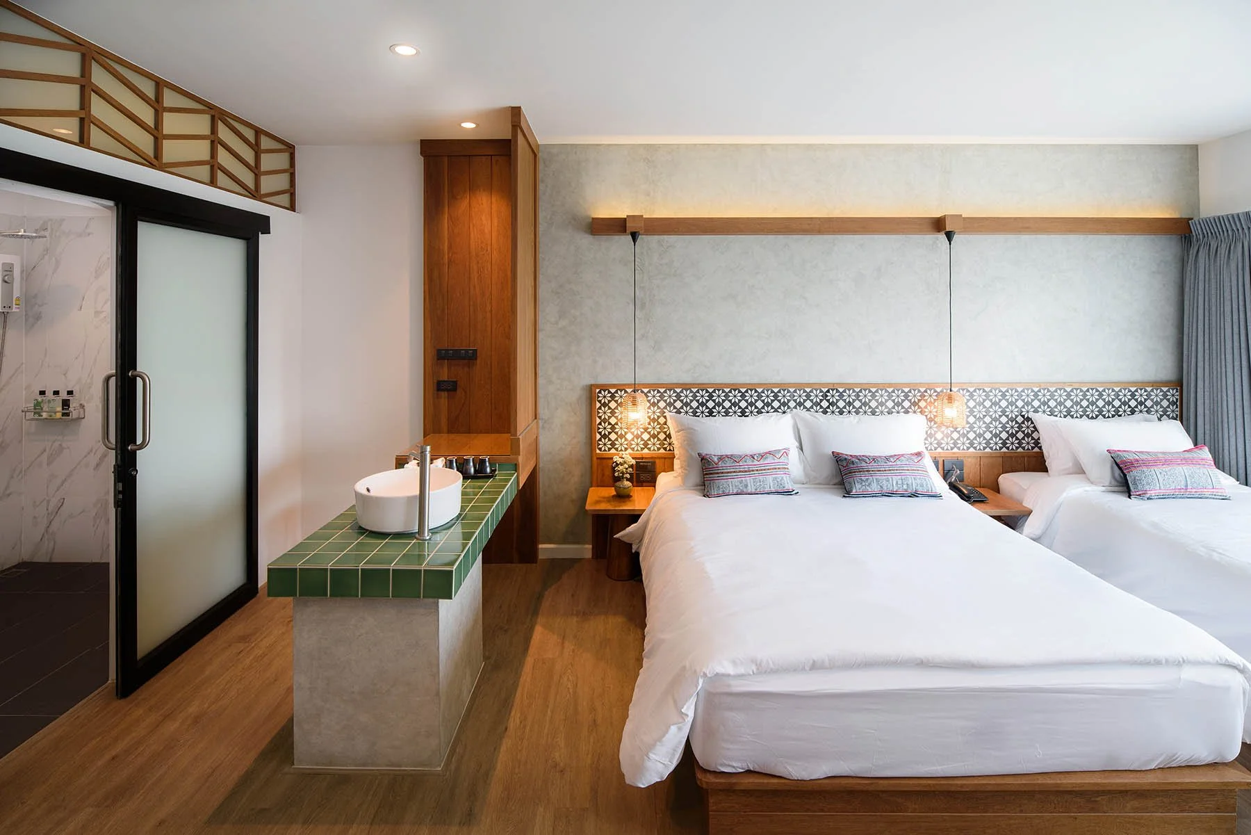 Hotel room with a large bed, two hanging pendant lights, a small bedside table with a vase, and a partially visible bathroom with a frosted glass shower door and a green-tiled sink.
