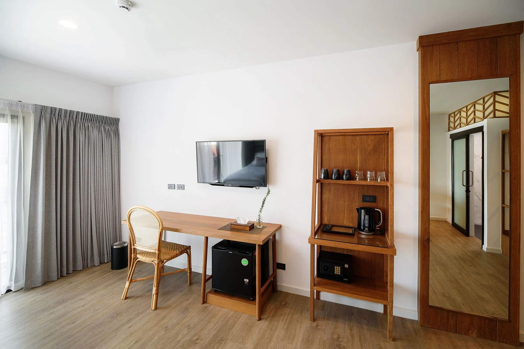 Hotel room with a wooden desk, a wicker chair, a flat-screen TV, a wooden shelf with glasses and a kettle, a safe, and a full-length mirror, with closed curtains and a sliding glass door.
