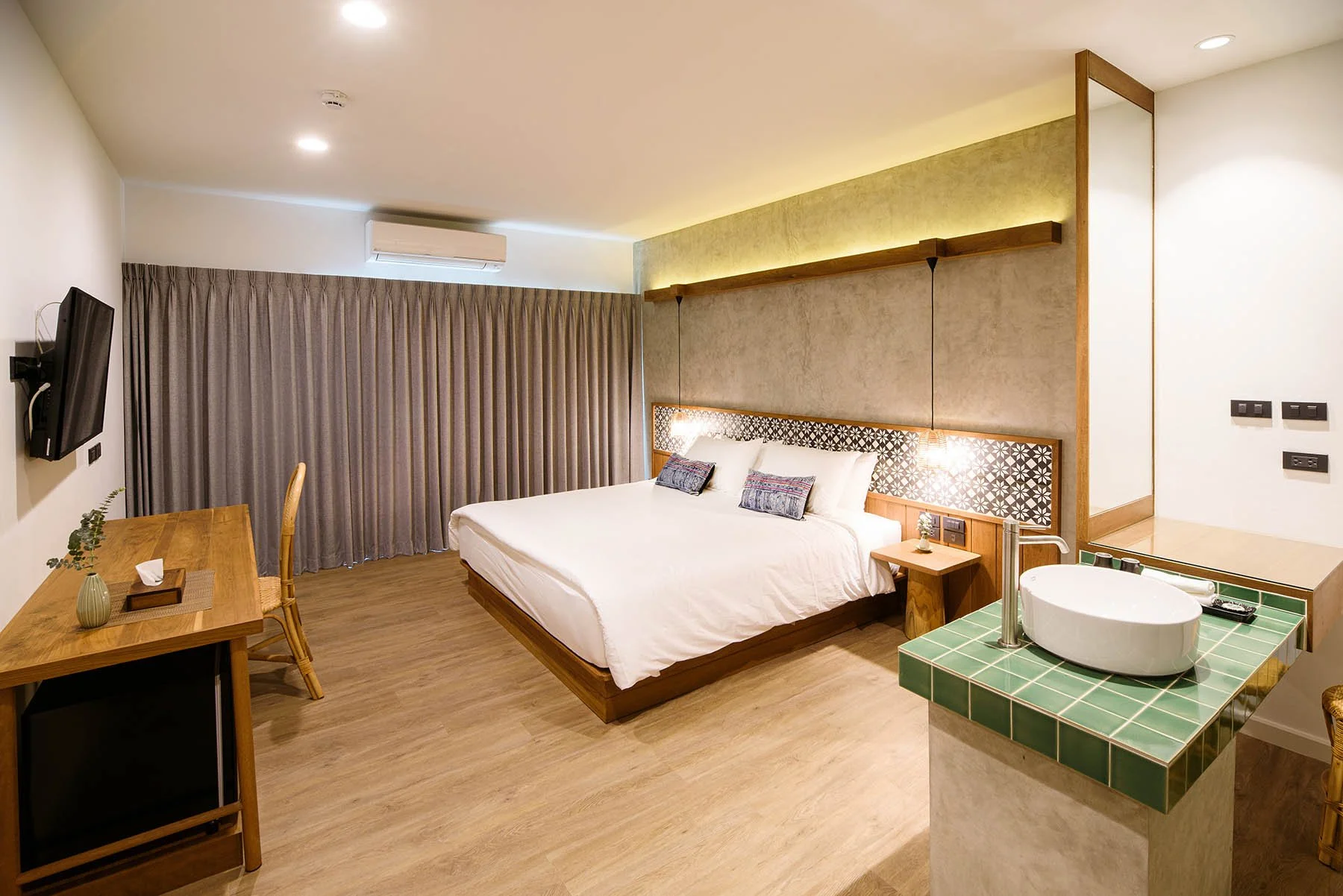 Hotel room with a large bed, a wooden desk with a chair, a wall-mounted TV, a sink with green tiles, and beige curtains.