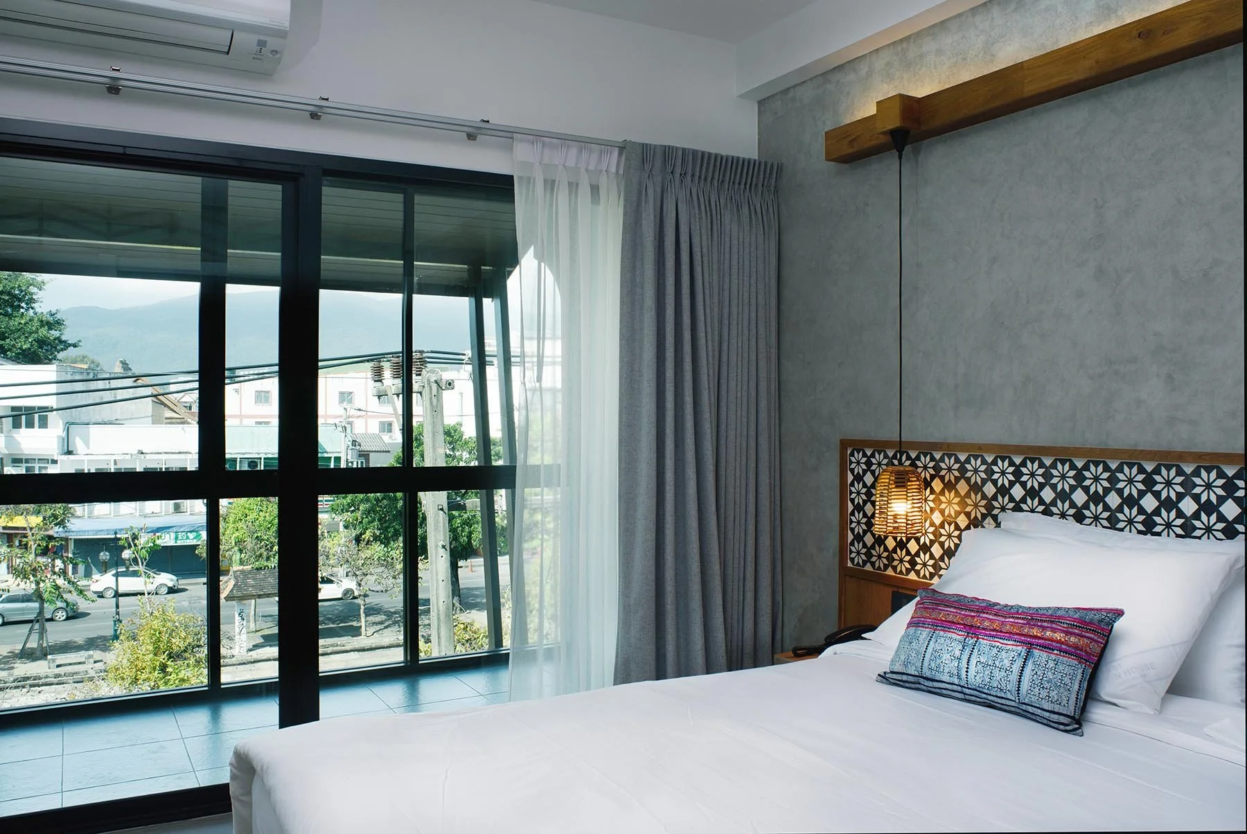 Hotel room with a bed, decorative pillow, hanging lamp, and large glass sliding door with curtains, overlooking a street and mountains outside.
