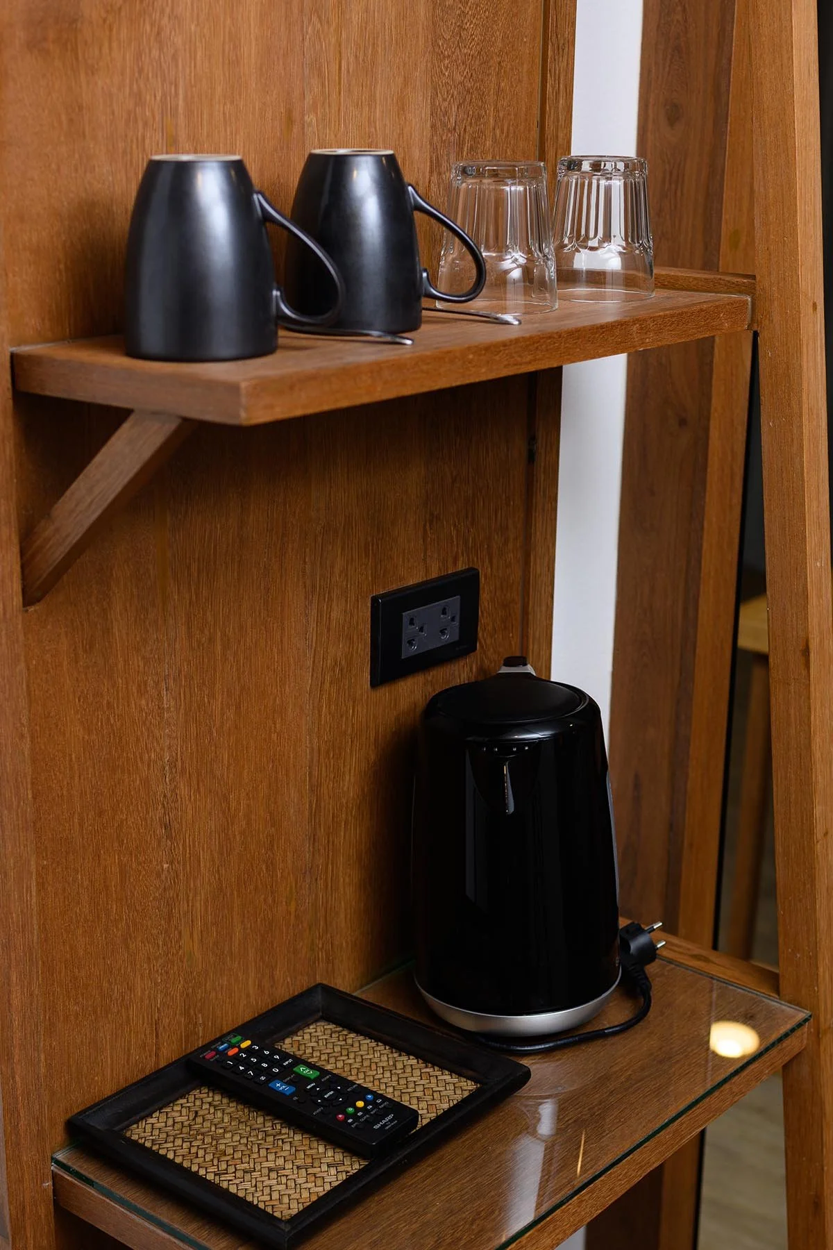 A wooden shelf unit with black coffee mugs, clear glasses, a black electric kettle, a landscape mirror, and a TV remote on a woven mat.