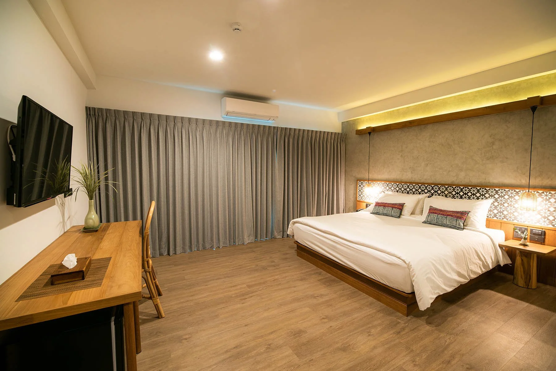 Hotel room with a large bed, wall-mounted TV, wooden desk with a chair, curtains, and warm lighting.