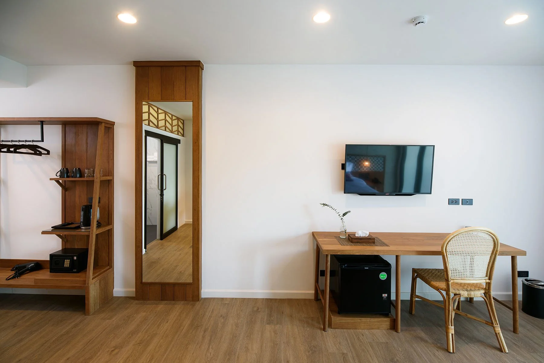 Modern hotel room with wooden furniture, flat-screen TV on the wall, mirror, small table with plant, and a wicker chair.