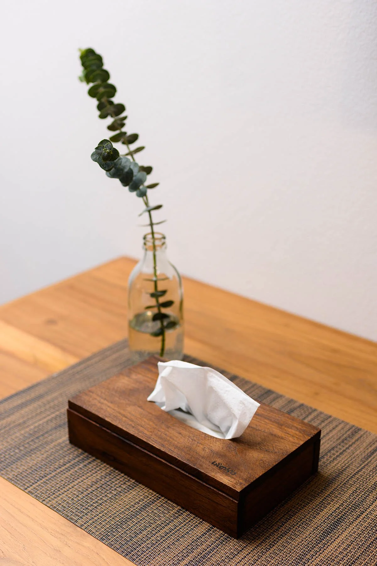 A wooden tissue box with a tissue protruding from it on a woven placemat on a wooden table, with a small glass vase holding a single stem of eucalyptus leaves in the background.
