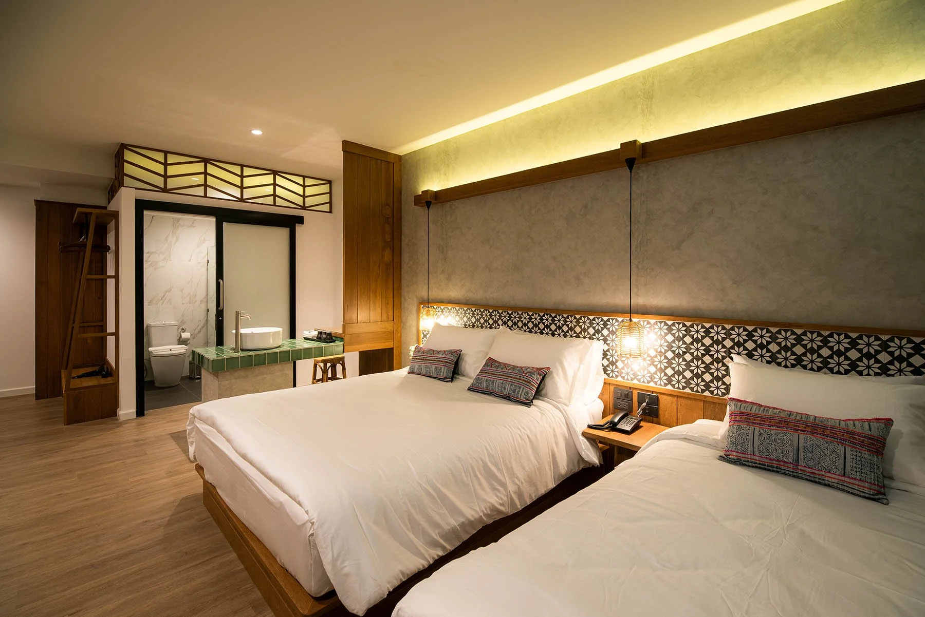 Hotel room with two beds, bedside table with phone, wall-mounted lamps, and bathroom with toilet and sink in the background.