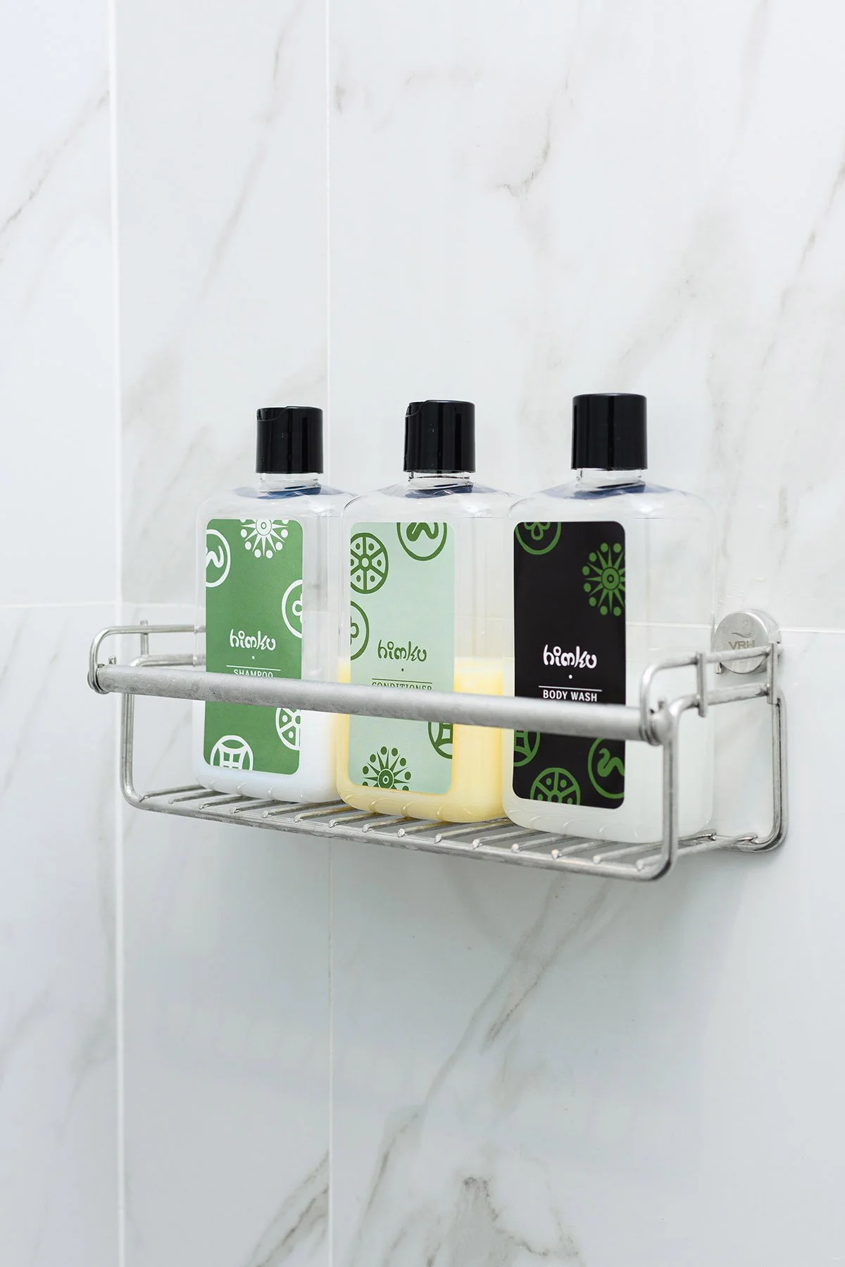 A metal shower shelf holding three bottles of shampoo, conditioner, and body wash mounted on a white marble tiled bathroom wall.