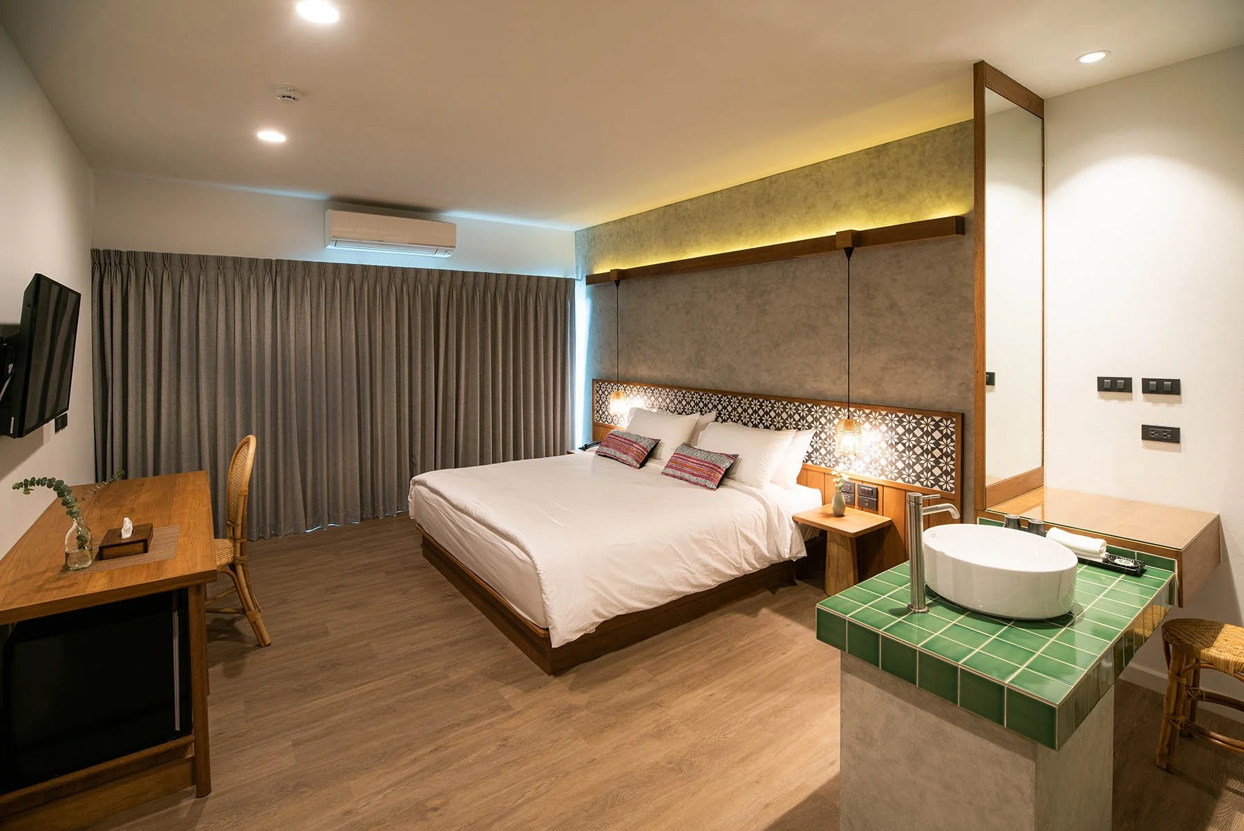 Modern hotel room with a bed, nightstand, wall-mounted TV, desk with a chair, and a green-tiled sink area.