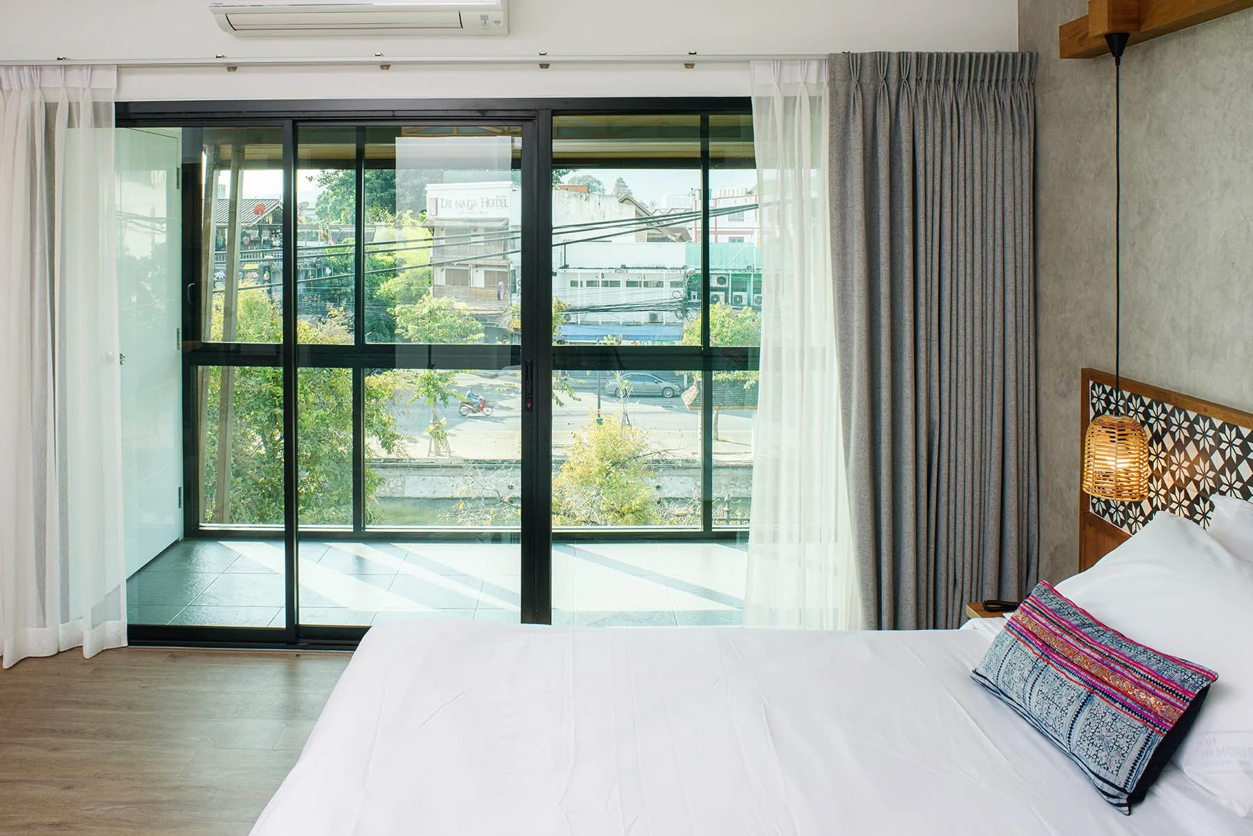 Hotel room with a large glass sliding door leading to a balcony, white curtains, a bed with white linens, and a decorative pillow, with city buildings visible outside.