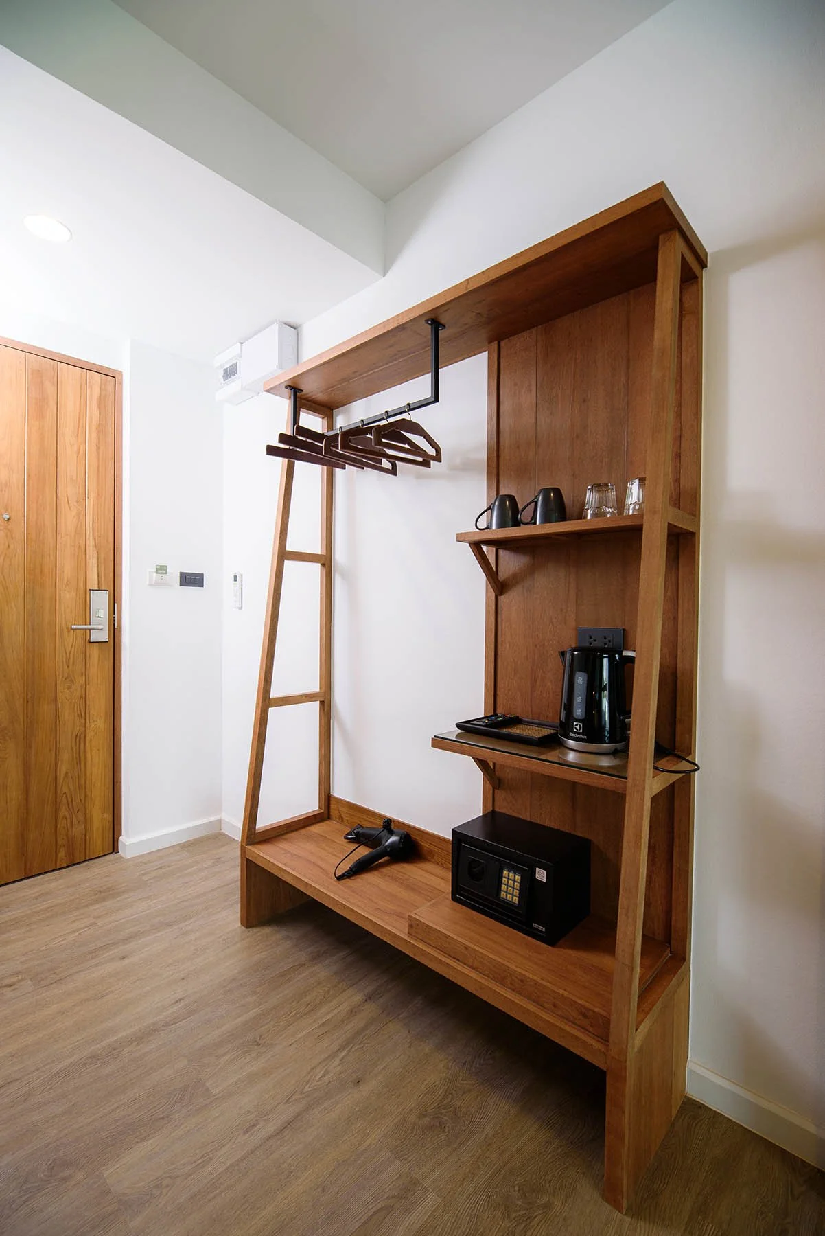 Wooden hotel room rack with hangers, shelves, a kettle, two cups, a remote, a hairdryer, and a small safe