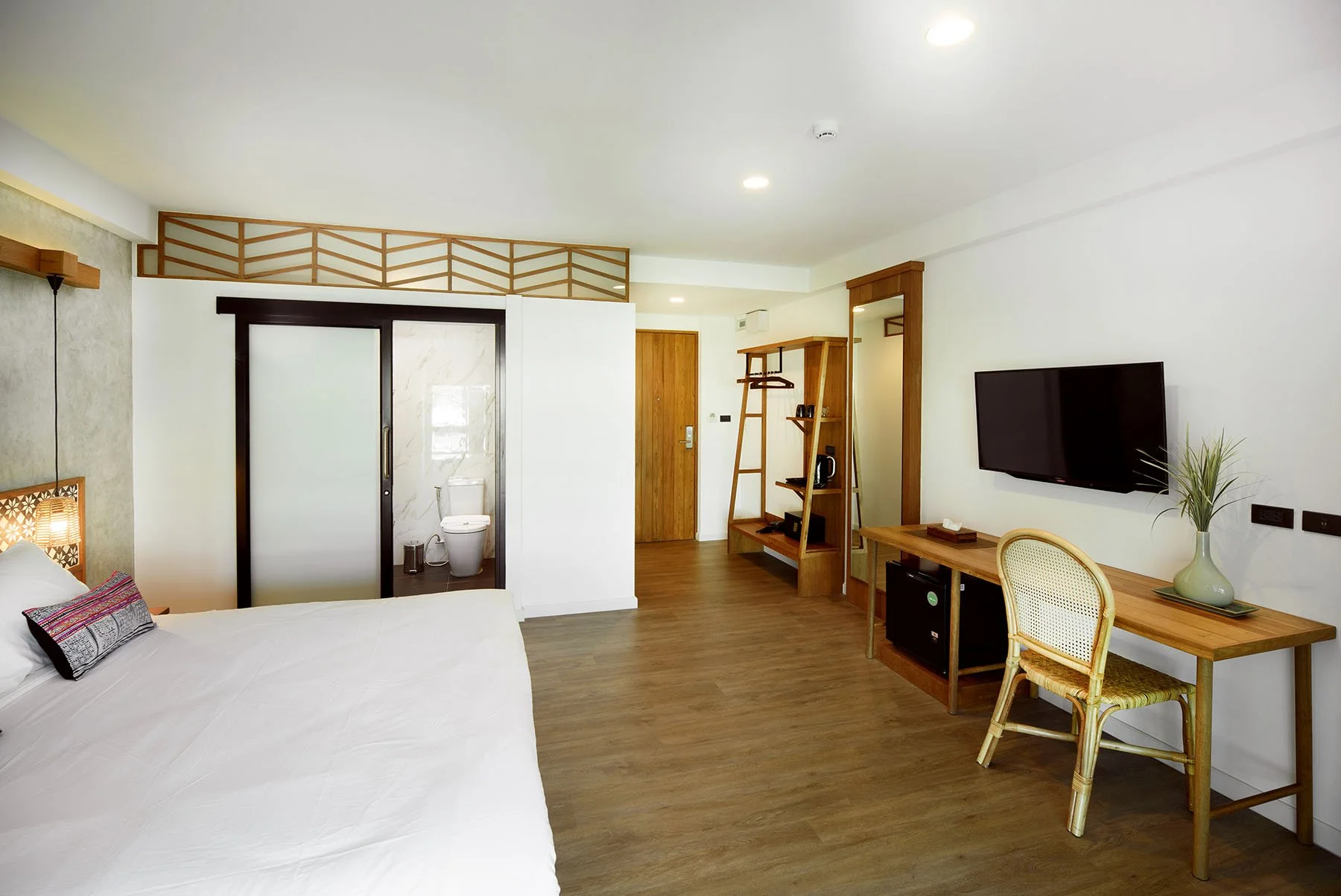A hotel room with a bed, a TV mounted on the wall, a wooden desk with a chair, a mini fridge, a unique shelving unit, and an open bathroom area with a toilet visible through sliding doors.