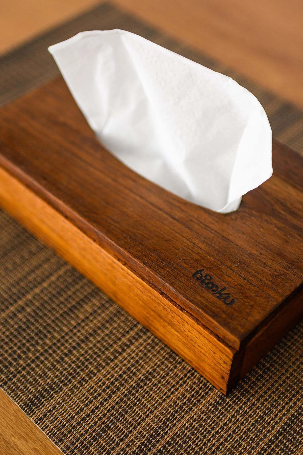 A wooden tissue box holder with a white tissue protruding from it, placed on a table with a woven placemat.