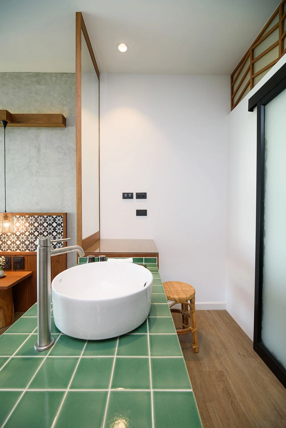 A modern bathroom with a green tiled countertop, a white vessel sink, and a stainless steel faucet. There is a small wicker stool and a frosted glass sliding door on the right. The background features a white wall with three black electrical outlets