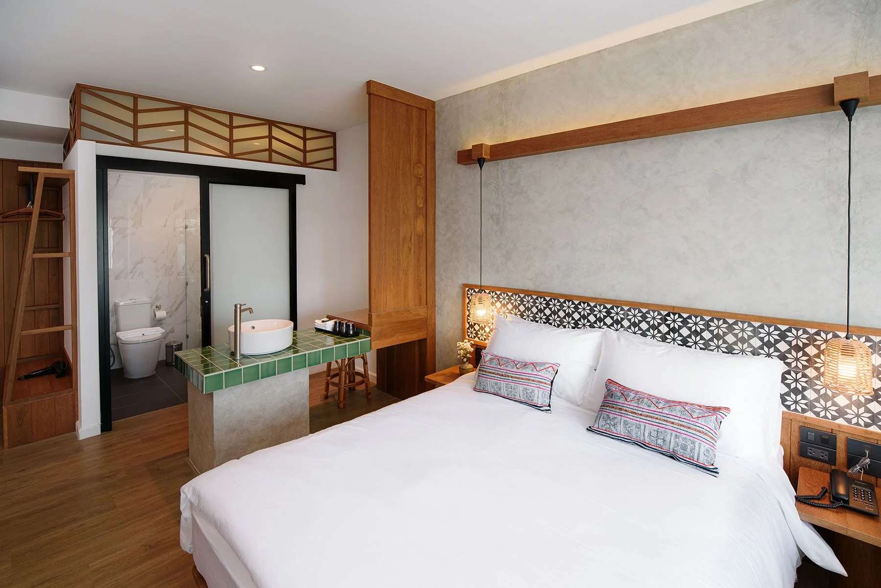 Modern hotel room featuring a white bed with patterned pillows, wooden accents, hanging bedside lamps, a small wooden nightstand with a telephone, and a bathroom with a toilet and a sink separated by a sliding glass door.