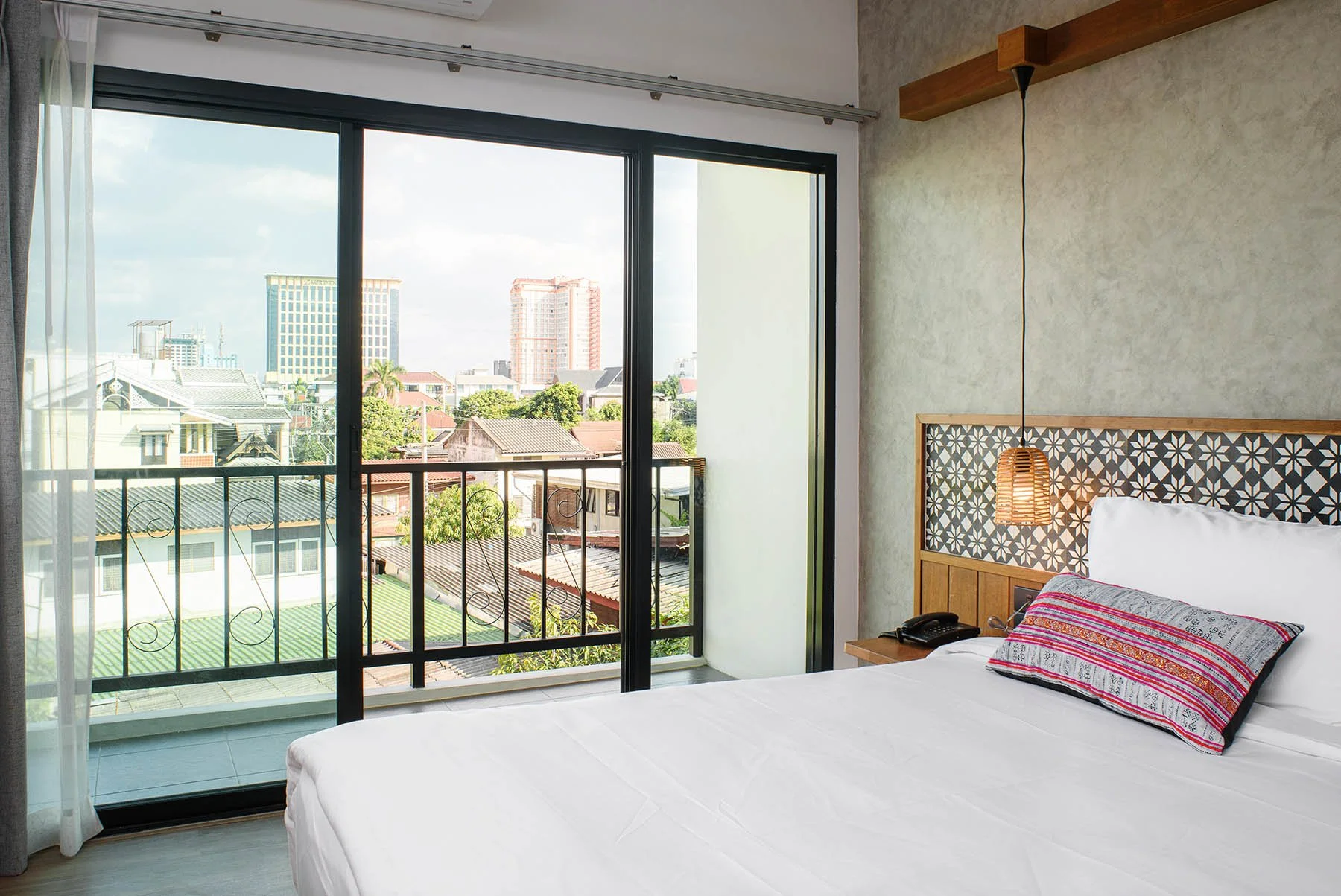 Hotel room with a bed, headboard, throw pillow, bedside phone, and a hanging lamp, overlooking a cityscape through a large sliding glass door leading to a balcony.
