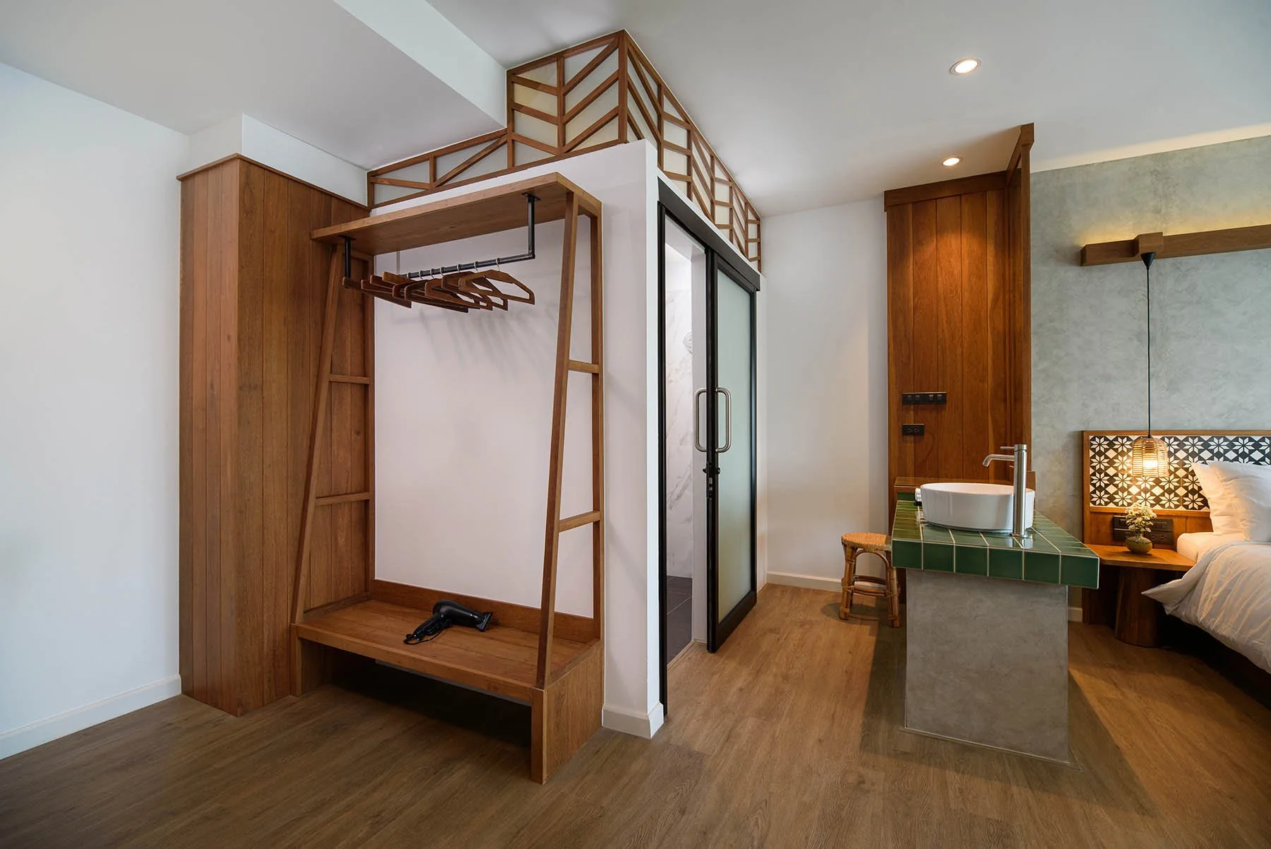 A hotel room with a wooden open closet, a hairdryer hanging, a glass shower door, a green tiled kitchen counter with a vessel sink, a small stool, a bed with white linens, and a wooden headboard with a decorative black and white pattern.