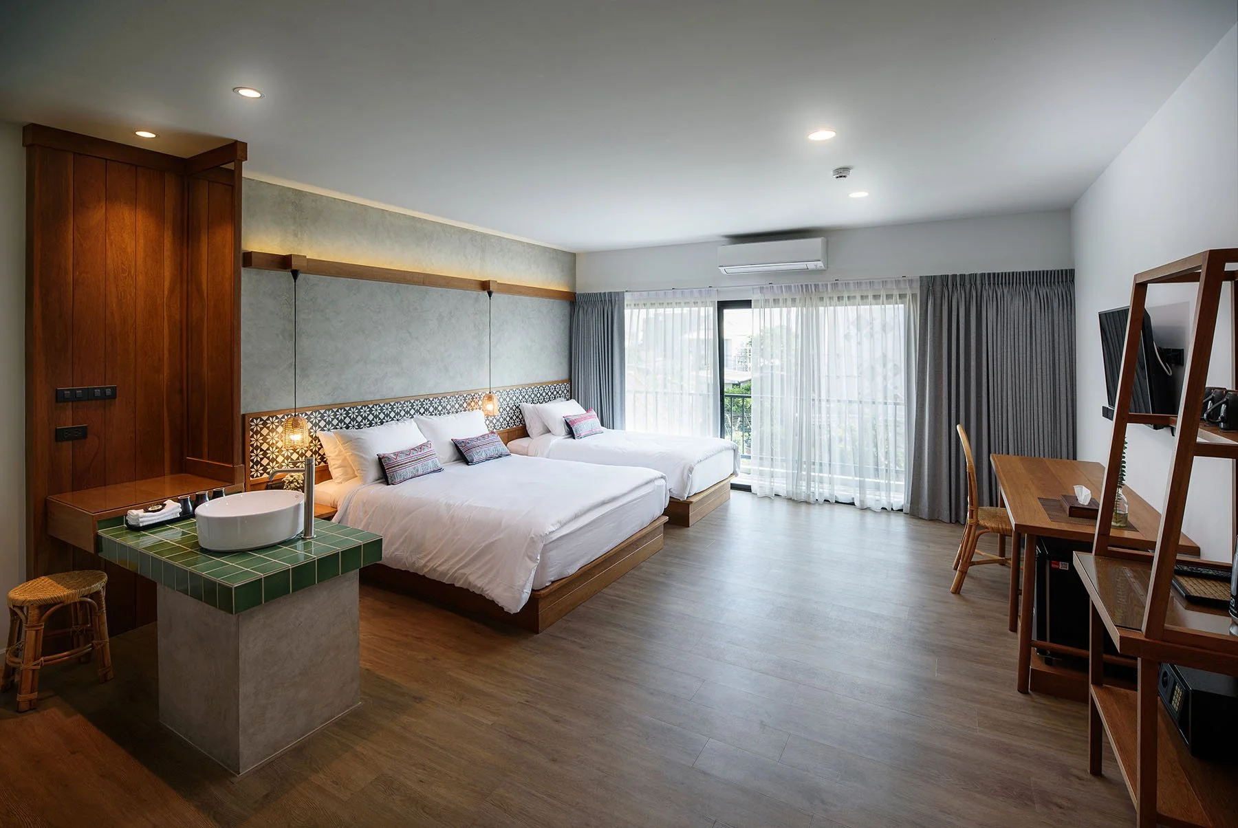 Modern hotel room with two beds, a balcony with curtains, wooden furniture, and a tiled sink area.