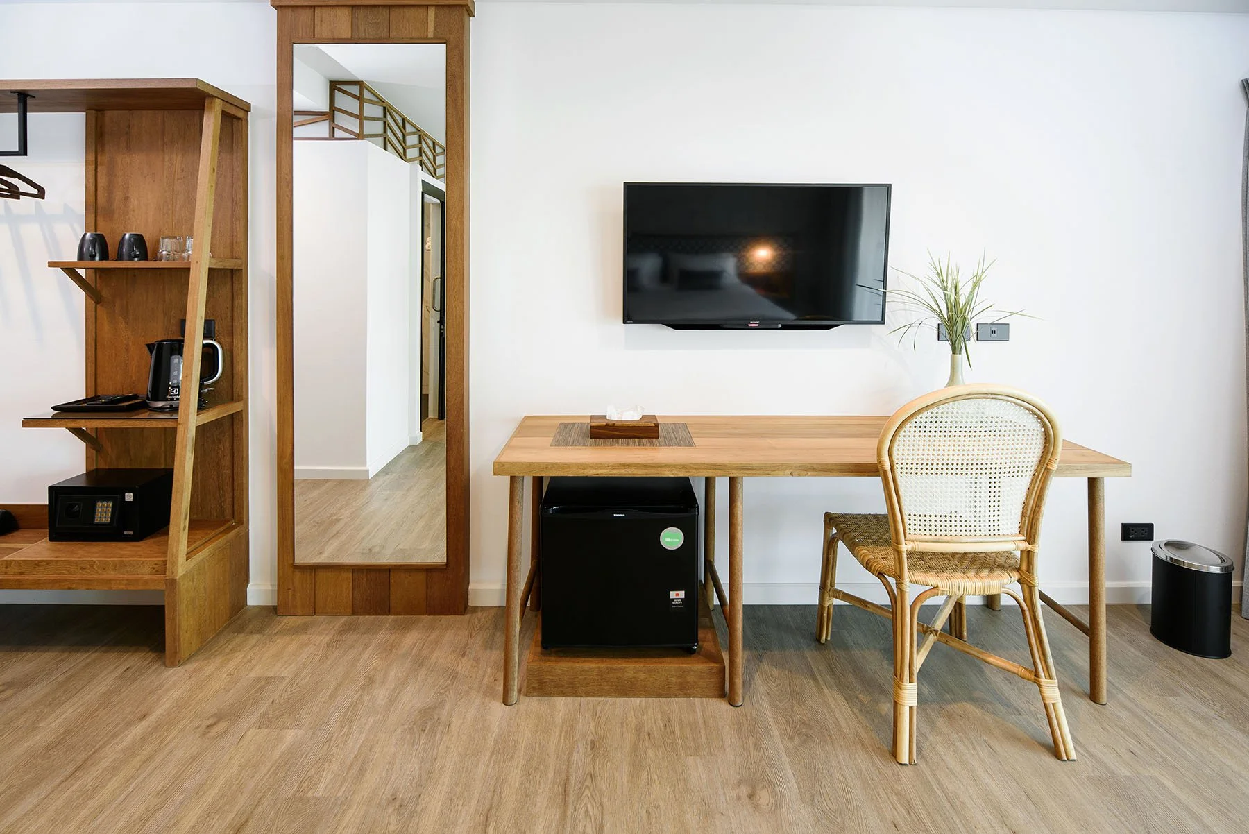 Interior of a room with a wooden desk, chair, a wall-mounted TV, a small fridge, a mirror, a wooden shelving unit with items, and a potted plant