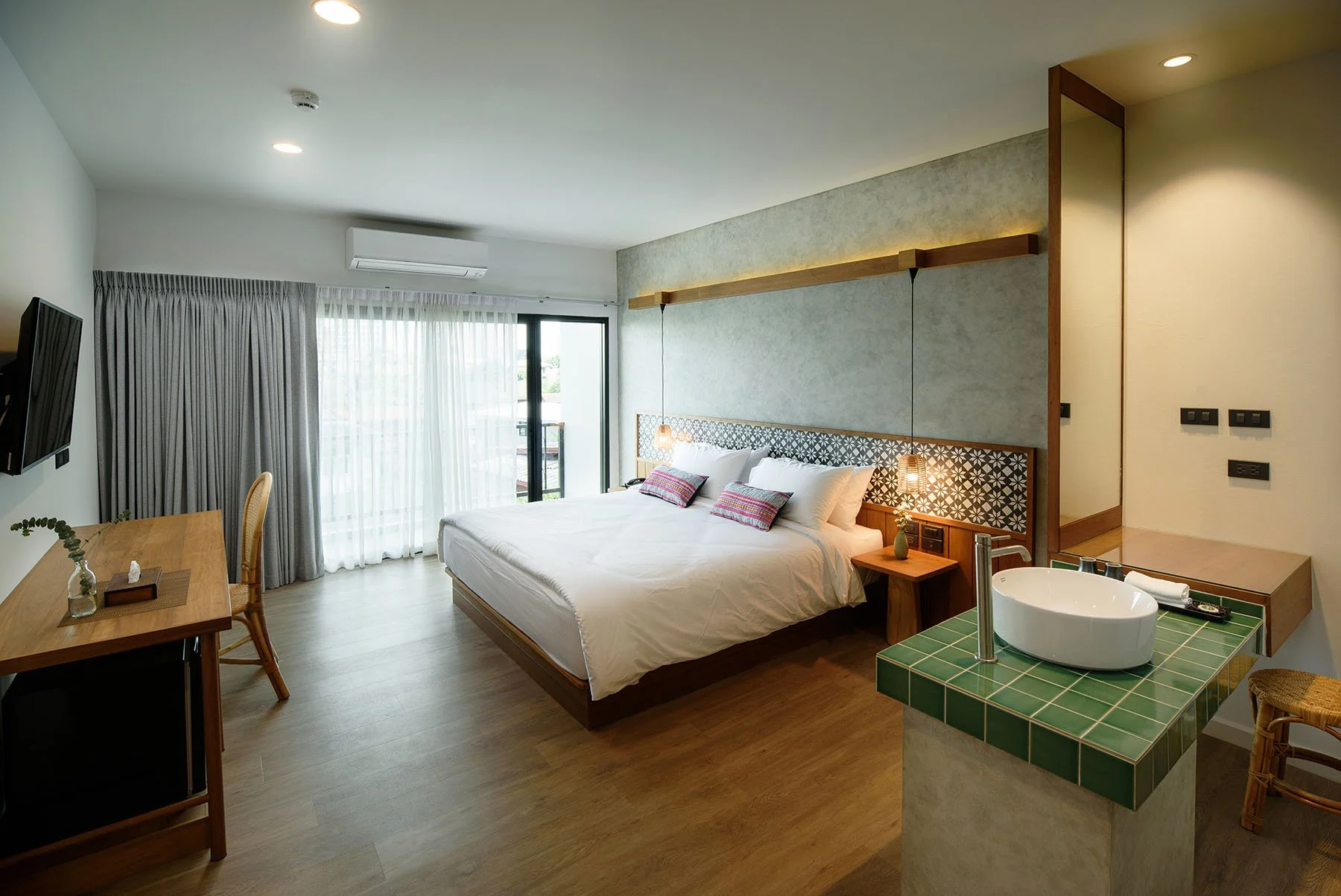 Hotel room with a large bed, bedside lamps, a desk, a chair, a seating area, and sliding glass doors with curtains leading to a balcony.