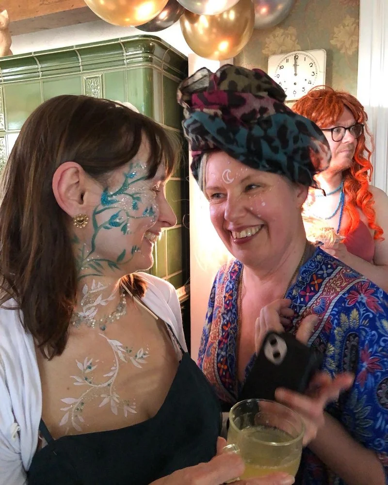 Naamiaisissa kasvomaalauksia, naisi anaurattaa ja taustalla parrakas mies Ariel peruukissaan mietteliäänä.