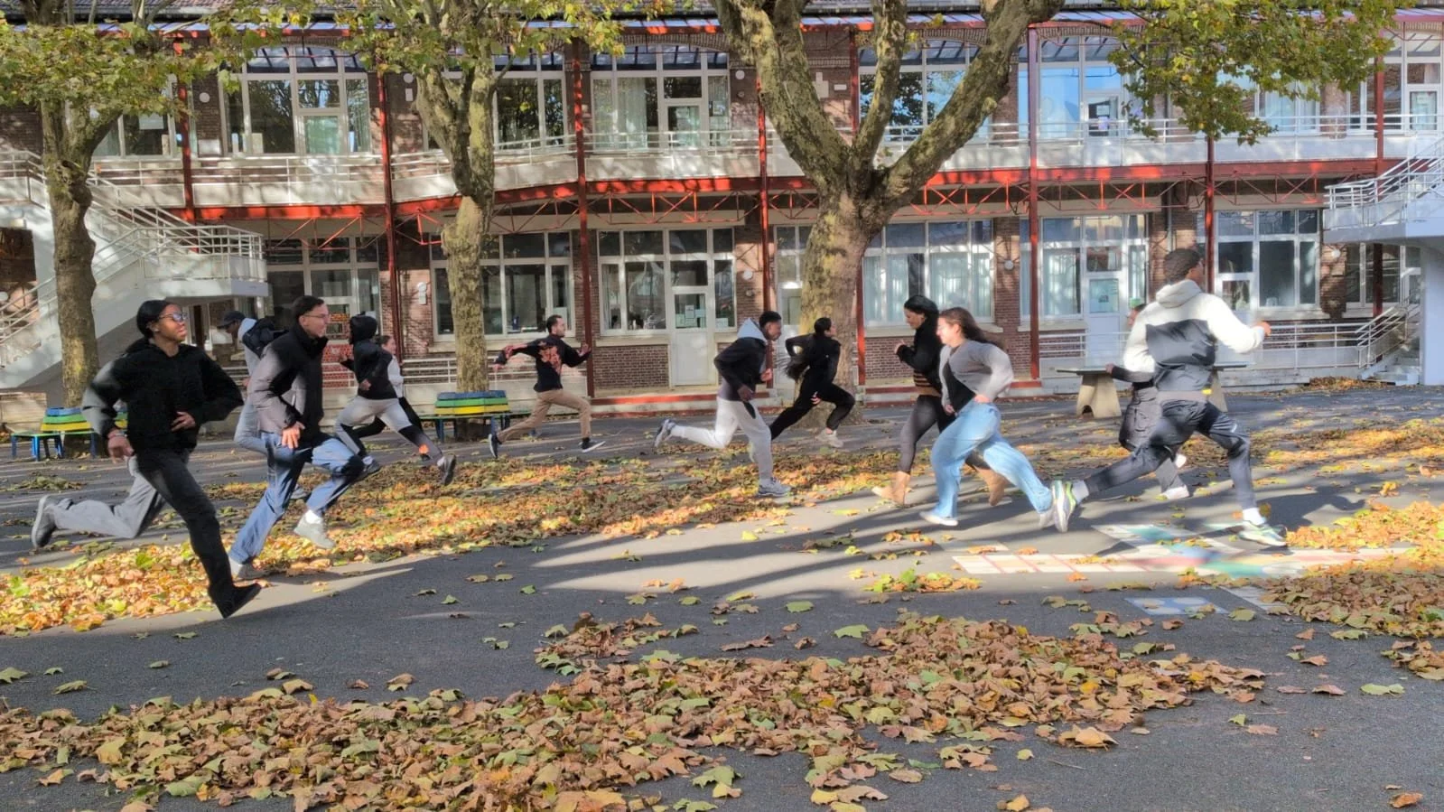 Groupe de personnes courrant à l'extérieur, avec des feuilles mortes sur le sol, sous un arbre en automne, devant un bâtiment moderne en briques et verre.