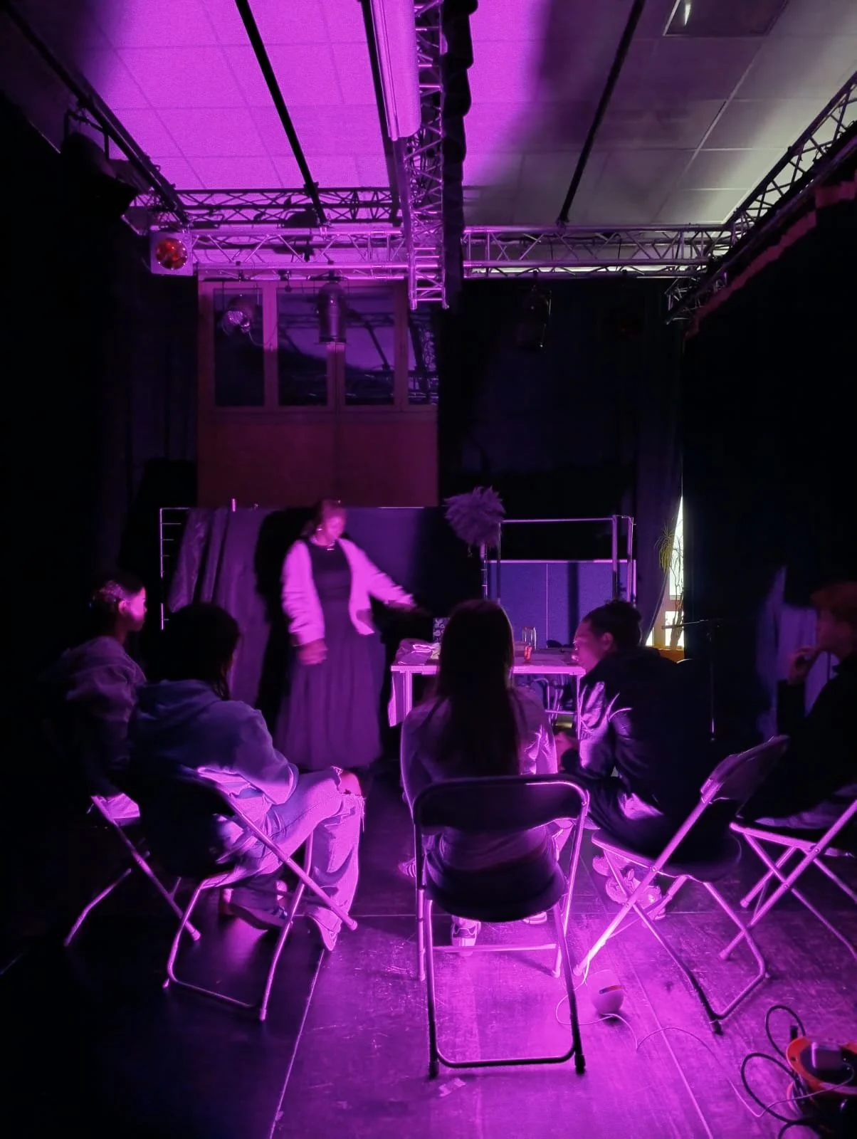 Une scène de théâtre ou spectacle avec des personnes assises devant une scène sombre et une femme debout, éclairée par des lumières violet et rose, dans une salle avec une structure en métal au plafond.