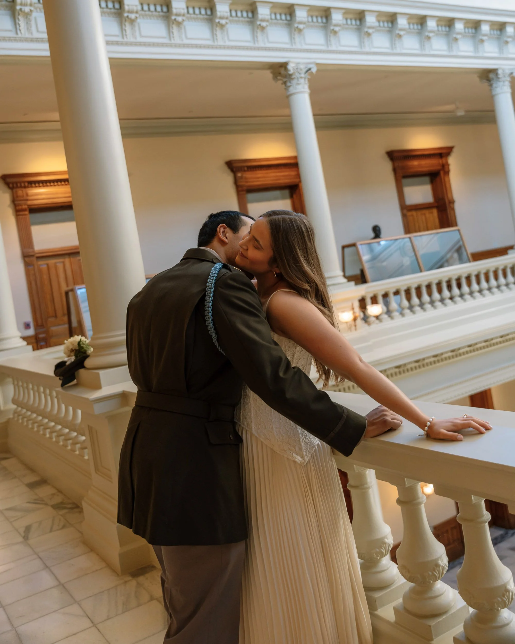 A couple sharing a kiss on a balcony inside a historic building with ornate columns and wooden accents.