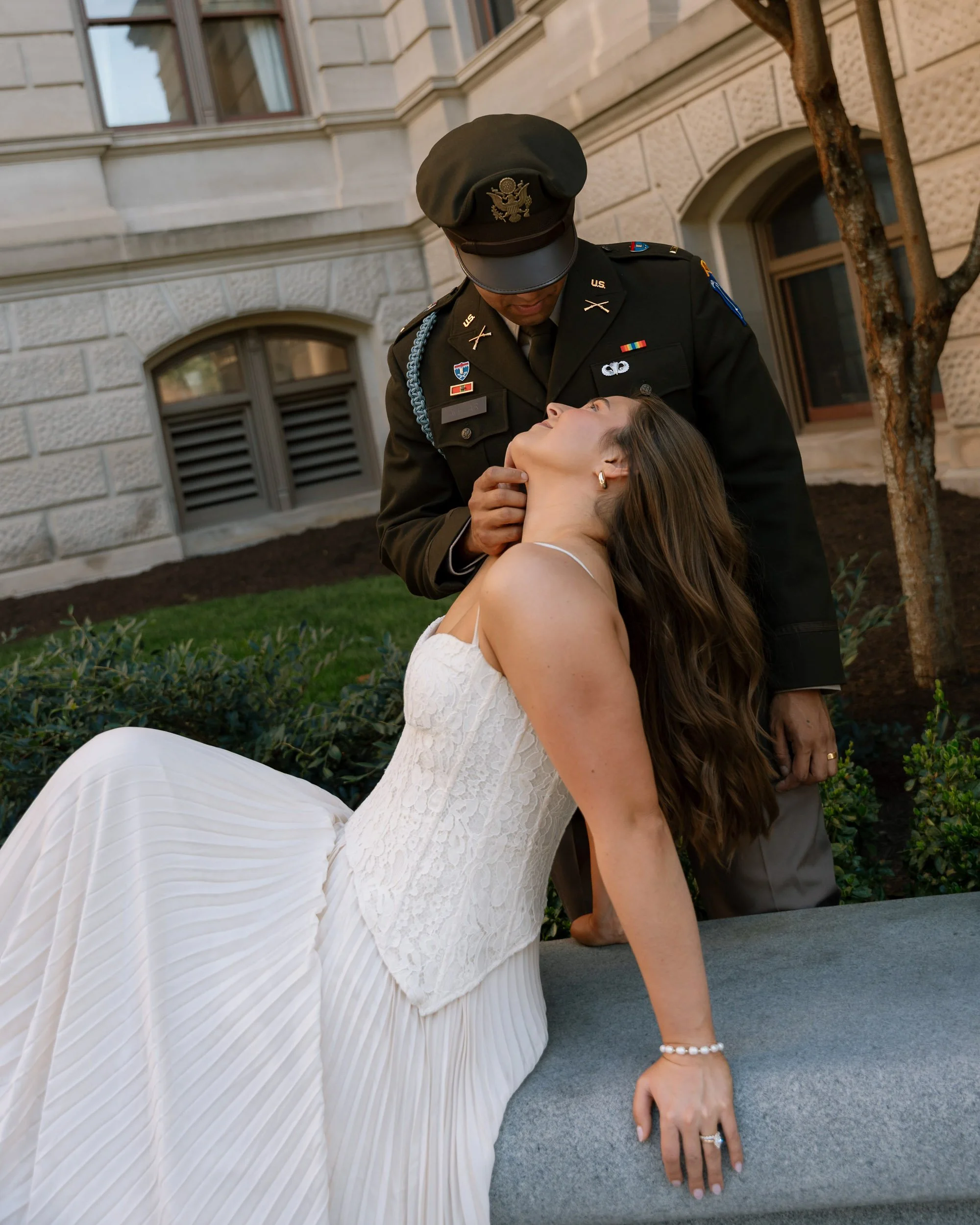 A woman in a white dress leaning back with closed eyes as a man in a military uniform gently touches her chin, outside a stone building with windows and a tree.