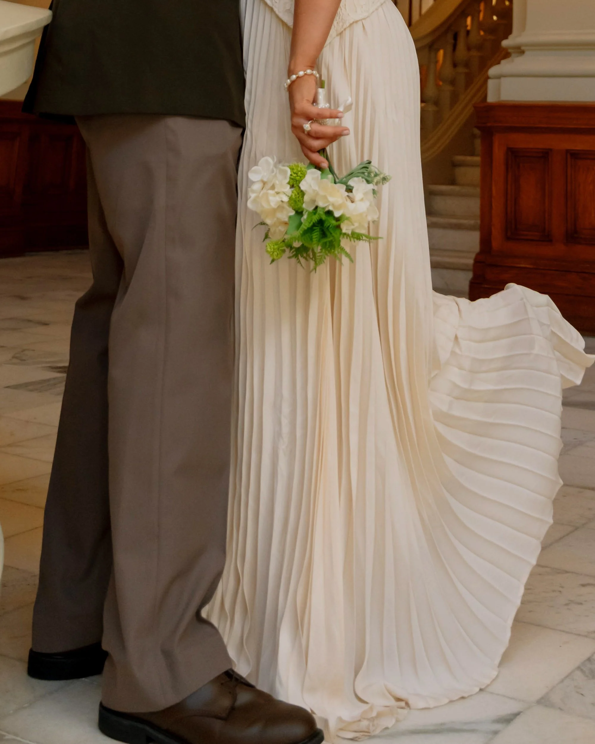 Bride and groom standing together, bride holding a bouquet of white and green flowers, bride wearing a long, pleated cream-colored dress, groom wearing gray pants and black shoes, staircase and wooden paneling in the background.