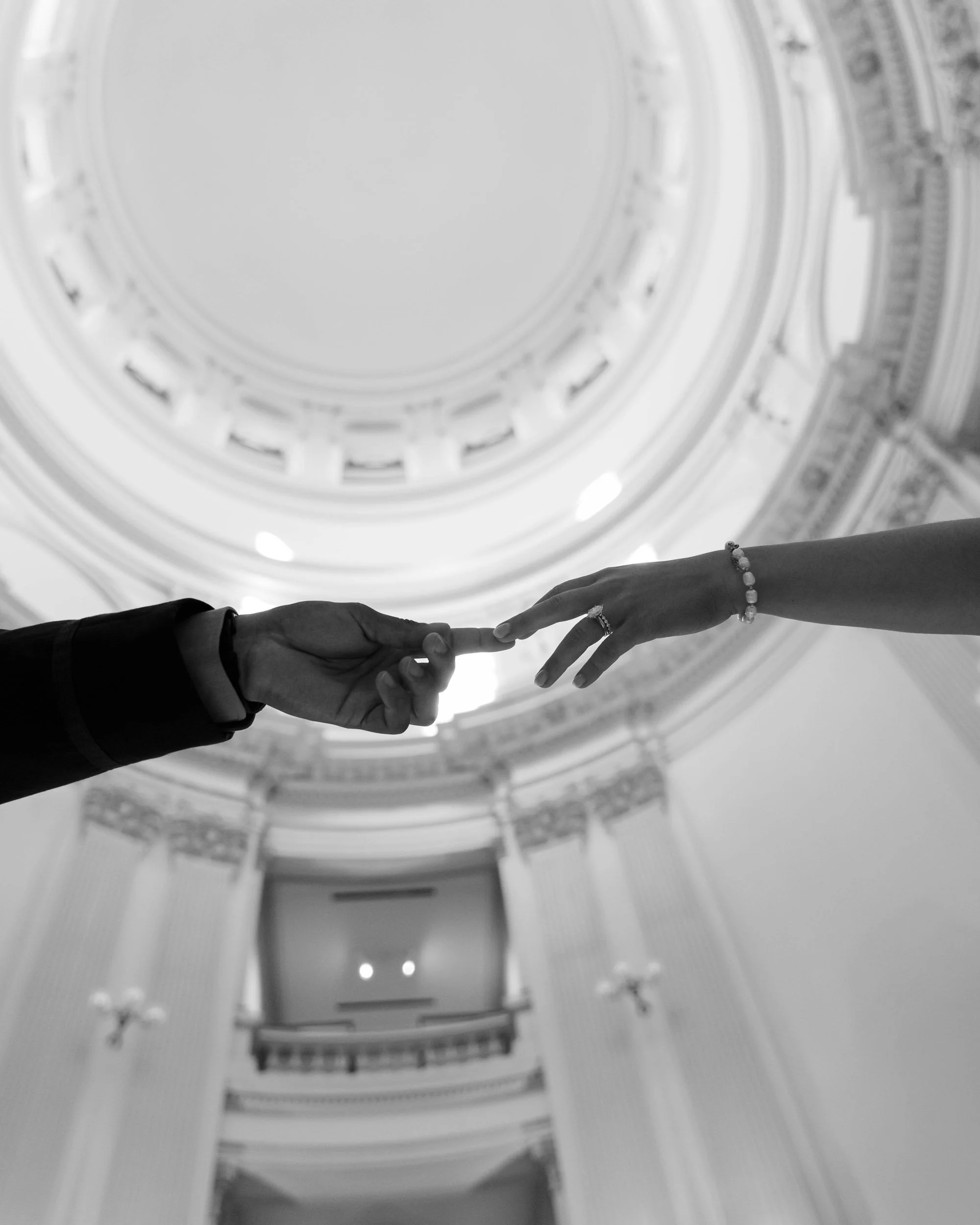 Black and white photo of a couple's hands reaching out towards each other, with the ceiling of an ornate building visible above.