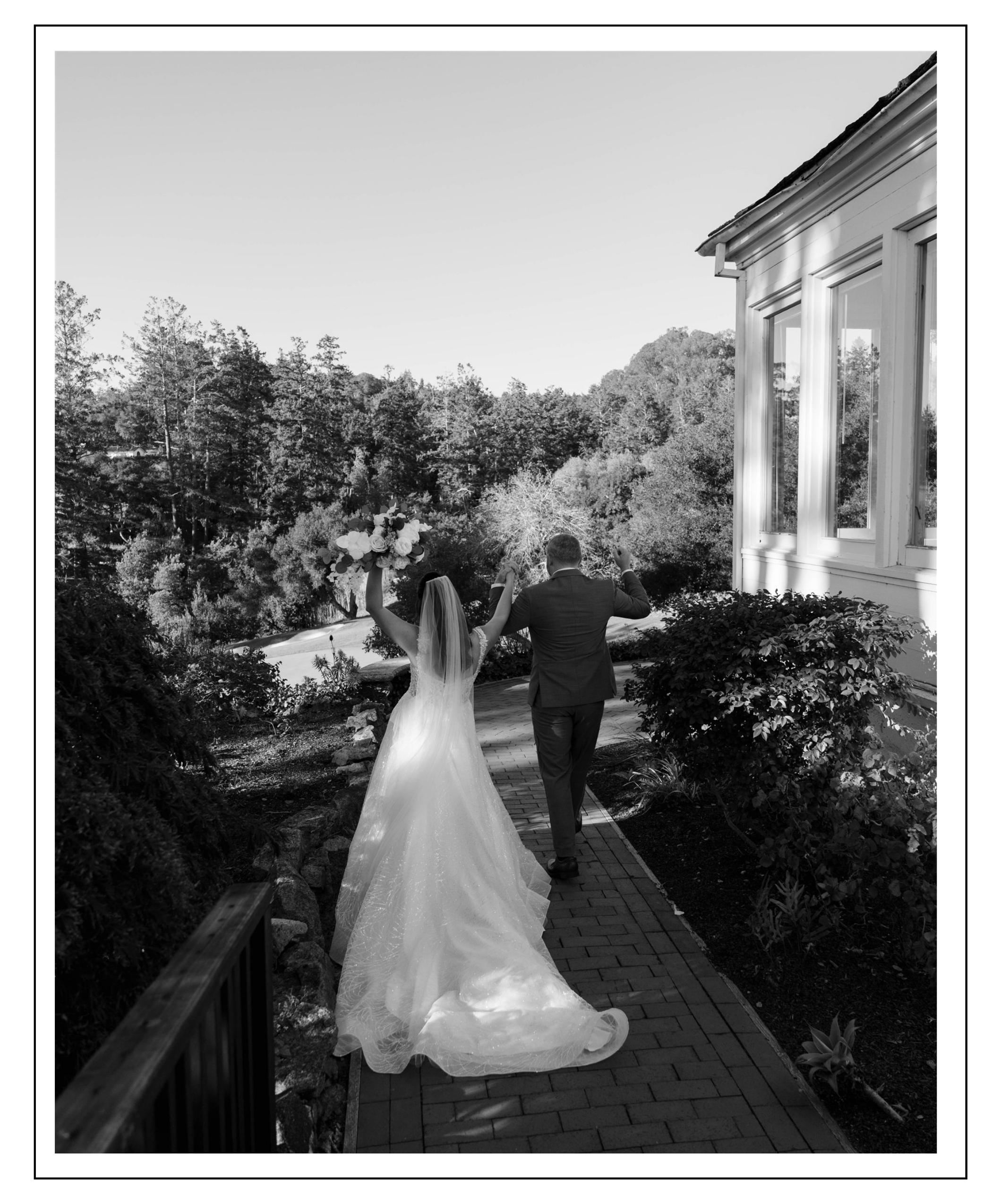 A black and white photo of a bride and groom walking away from the camera on a brick pathway, holding hands, with the bride holding a large bouquet and both raising their arms in celebration. The scene is outdoors near a house with large windows, surrounded by trees.
