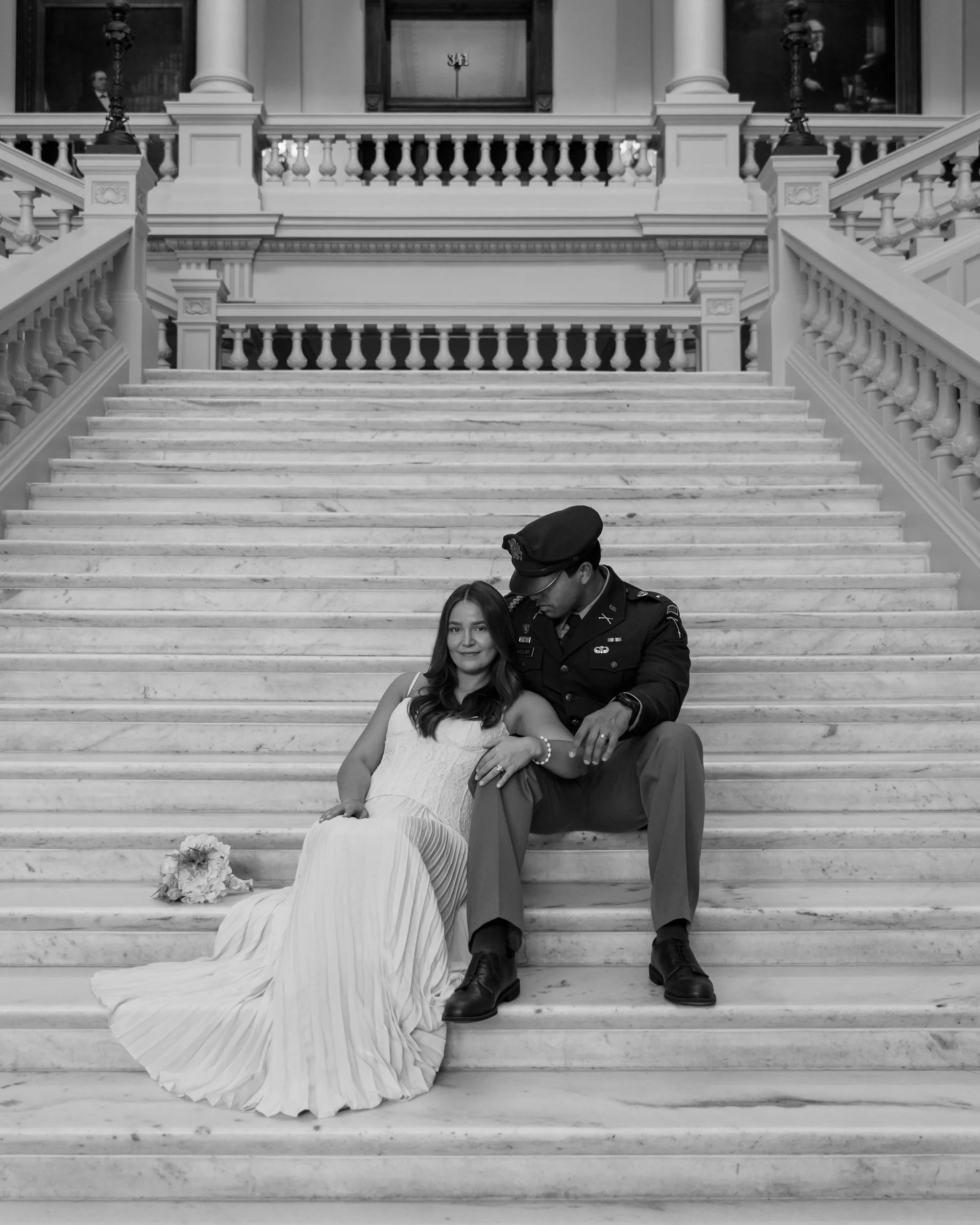 A woman in a white wedding dress sitting on marble stairs, with a man in military uniform sitting beside her, holding her arm. They are in a grand building with ornate columns and railings.