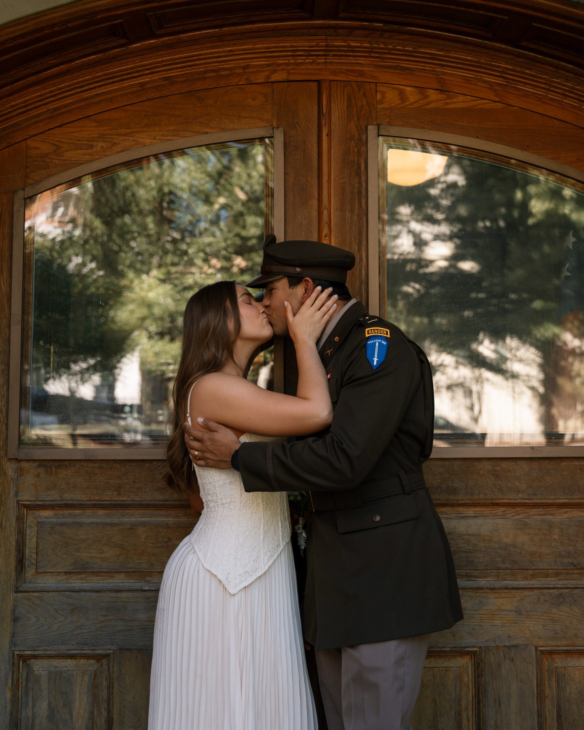 Couple kissing, woman in a white wedding dress and man in a military uniform, standing in front of a wooden door with glass panels, outdoors.