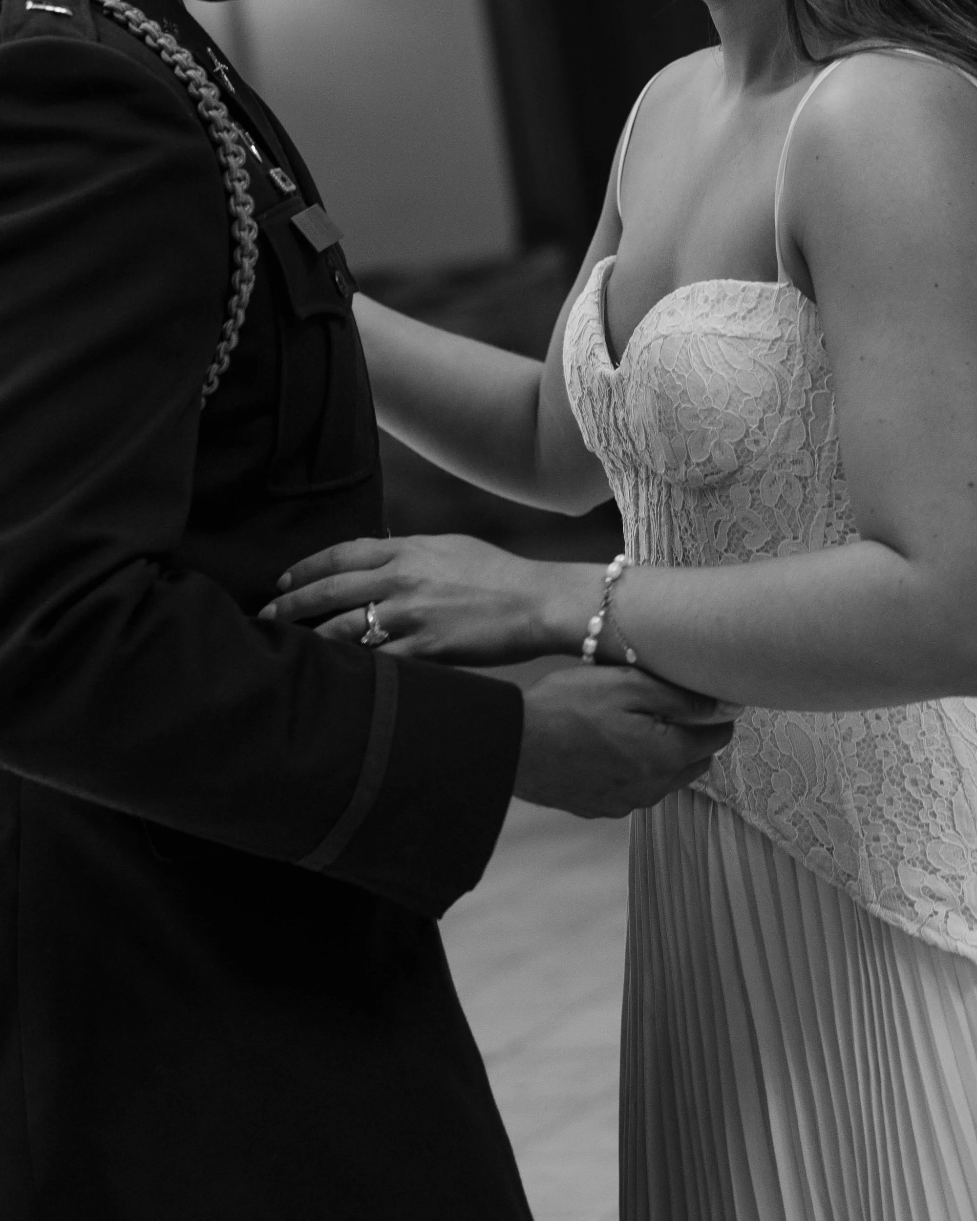 A black and white photo of a couple holding hands, the woman wearing a lace dress and jewelry, the man's attire including a jacket with a chain accessory.