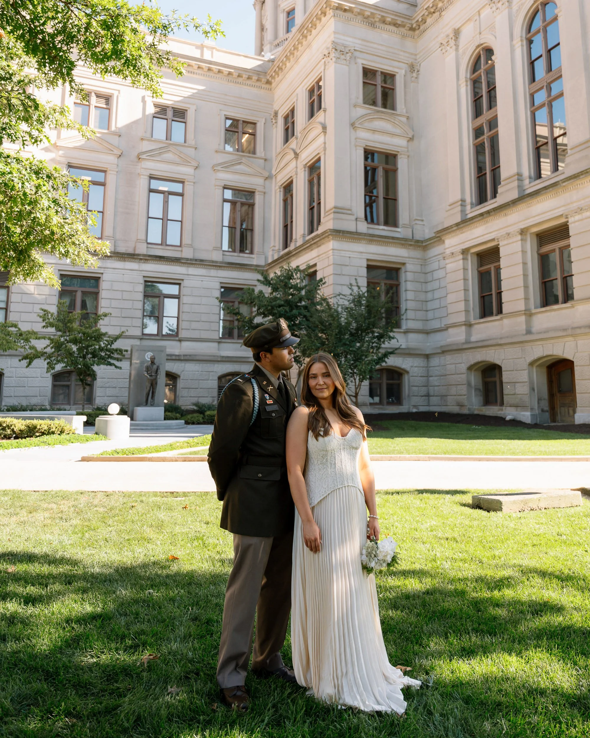 A woman in a white dress holding a bouquet stands next to a man in a military uniform on a grassy area in front of a large, historic building with tall windows and ornate architectural details.