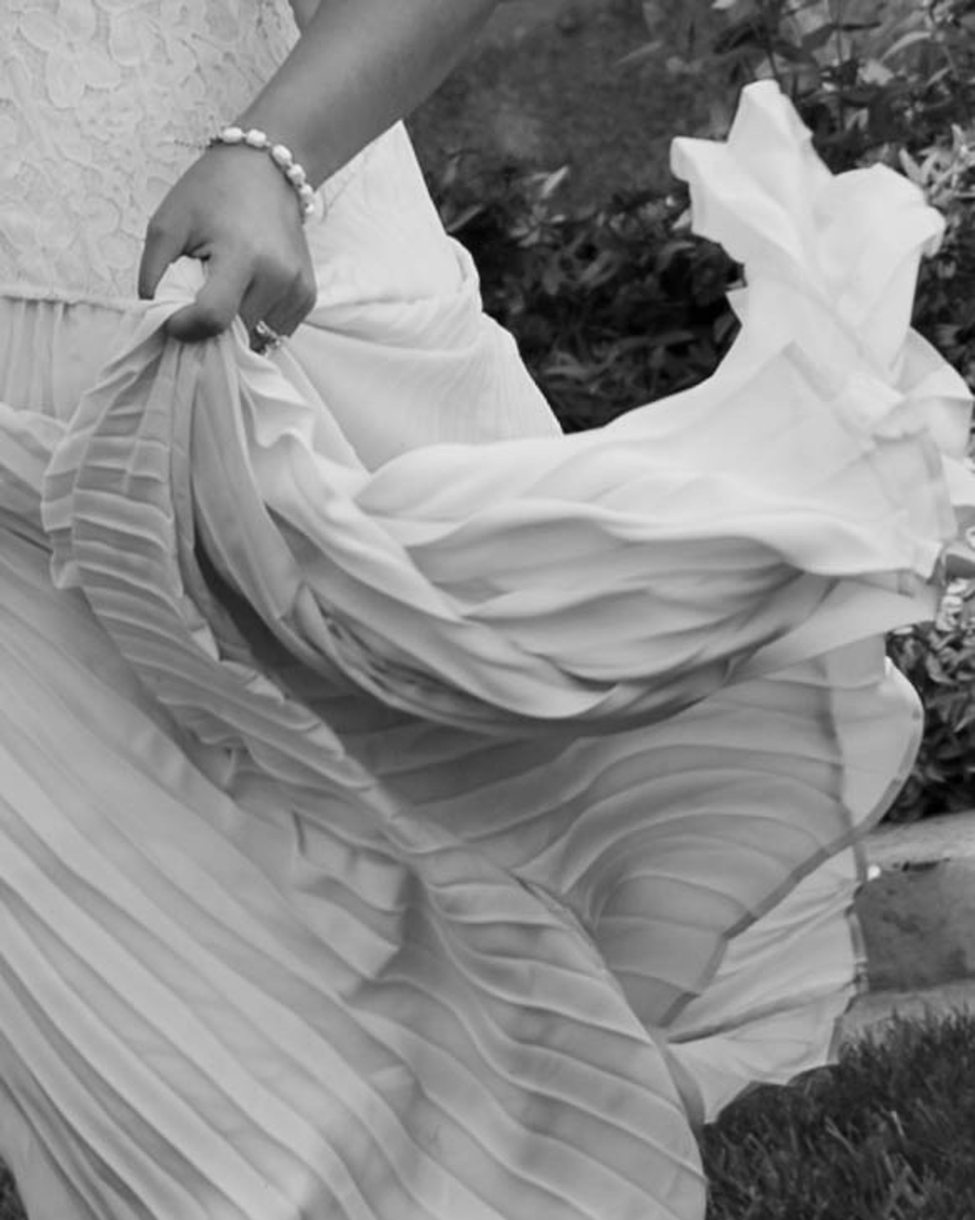 A person holding a flowing, pleated cloth or dress outdoors with grass and plants visible in the background.