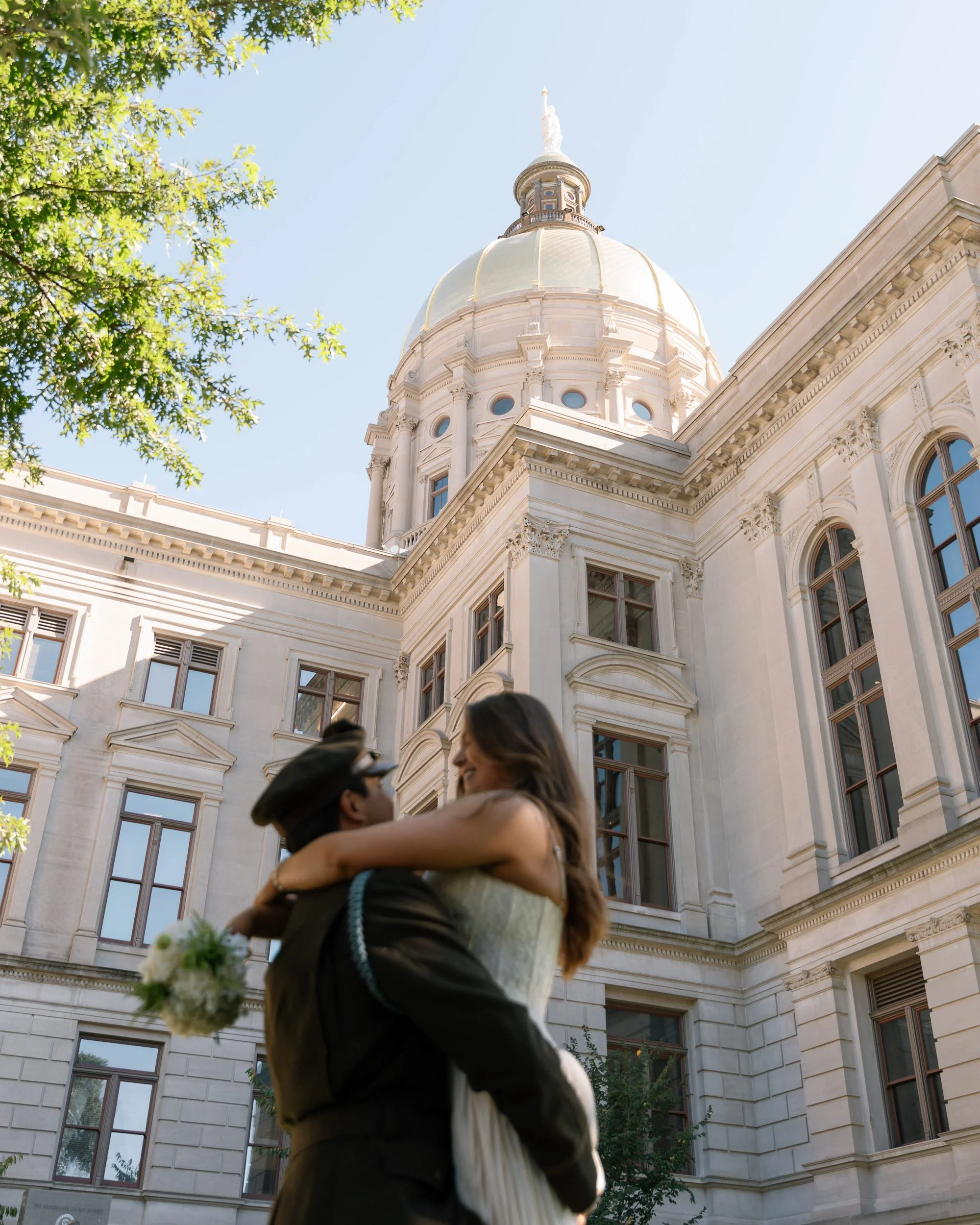 A couple, with the woman in a wedding dress and the man in a police uniform, embracing in front of a historic government building with a large dome and ornate architecture, under a clear blue sky.