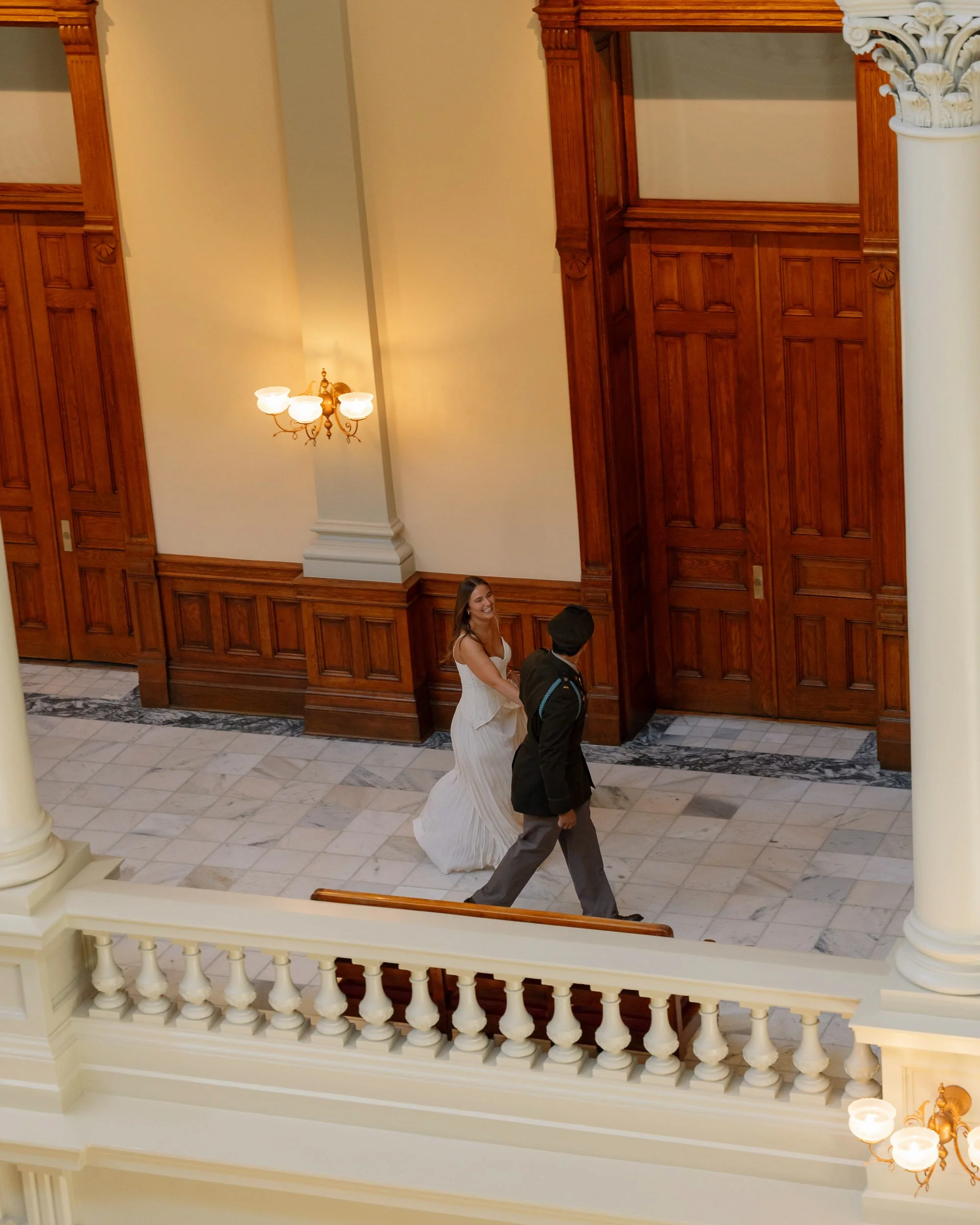 A bride in a white wedding dress walking with a groom in a black suit and hat inside a grand, wood-paneled hall with marble flooring and chandelier lighting.