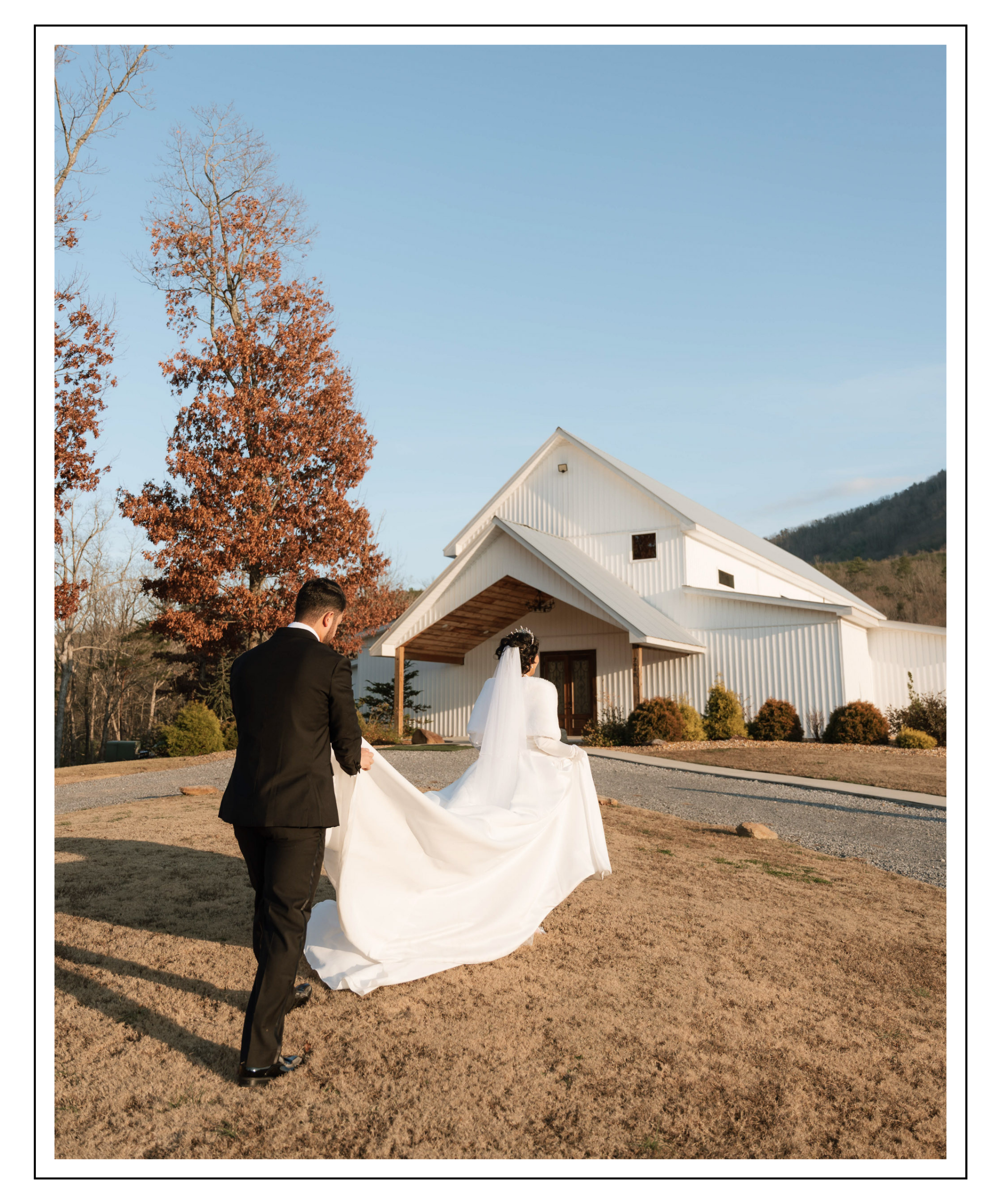 A bride and groom, the groom in a black suit and the bride in a white wedding dress, walking away from the camera toward a white barn-style building on a sunny day with a clear blue sky.