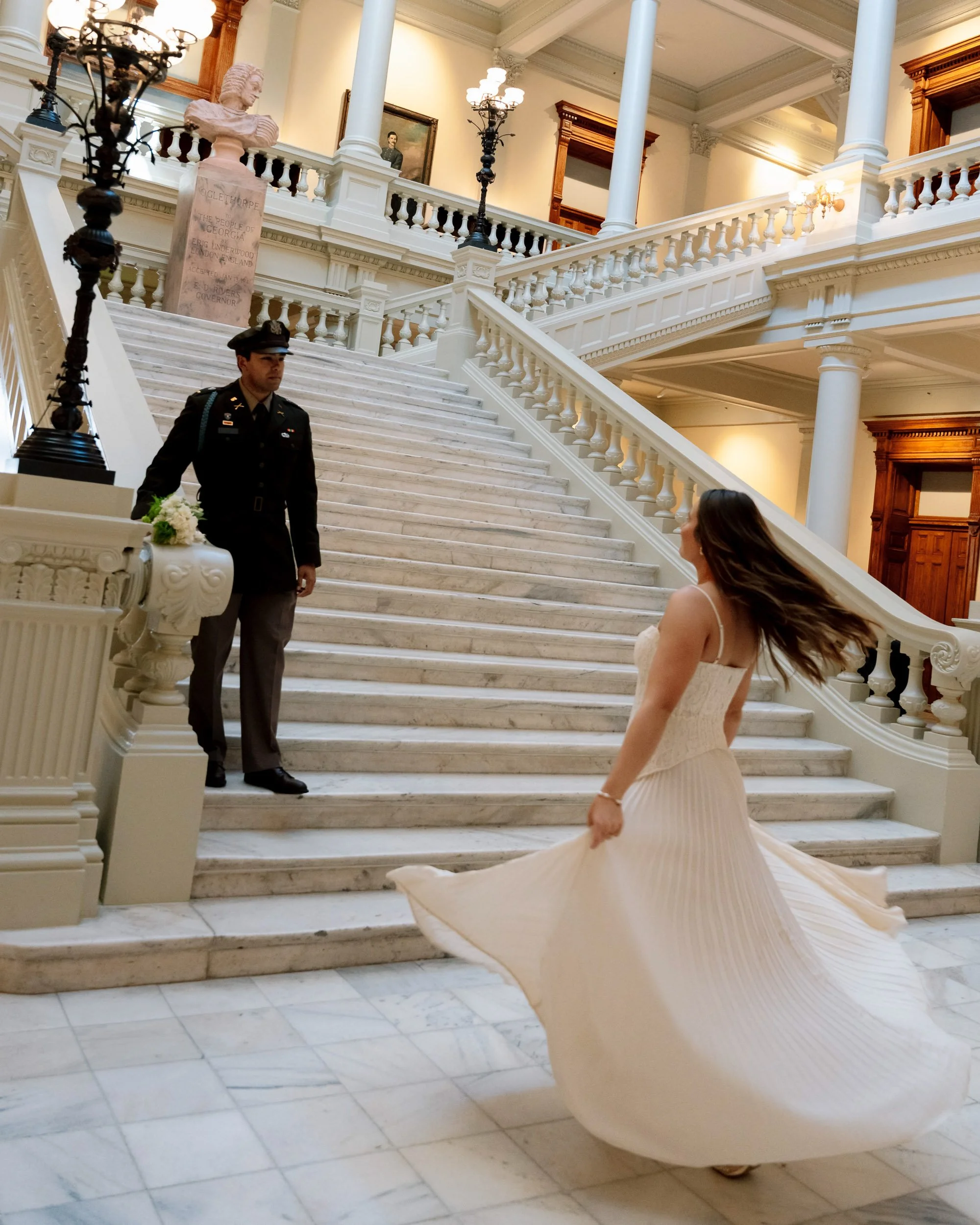 A woman in a white dress twirls on marble stairs with a guard standing at the bottom of the stairs inside a historic building.