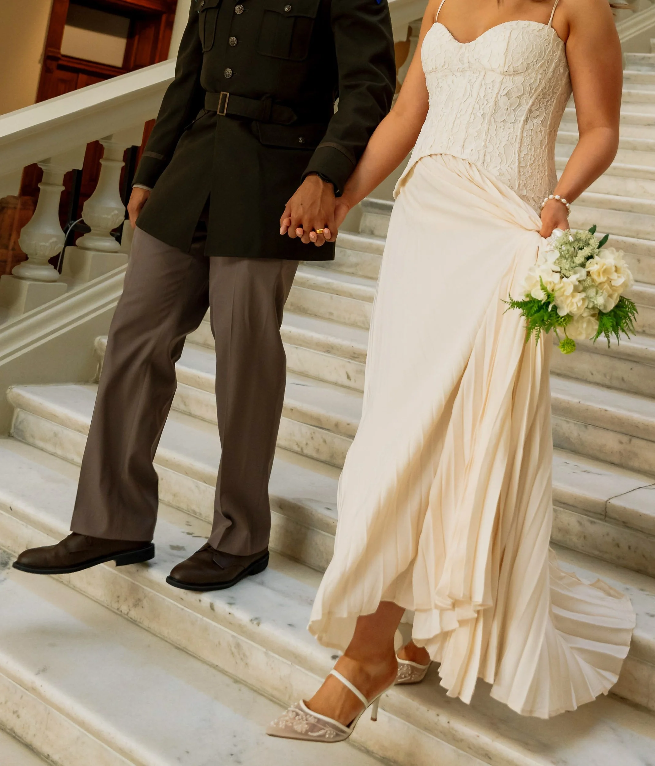Bride in a light-colored dress holding a bouquet, holding hands with a groom in a dark jacket, standing on marble steps.