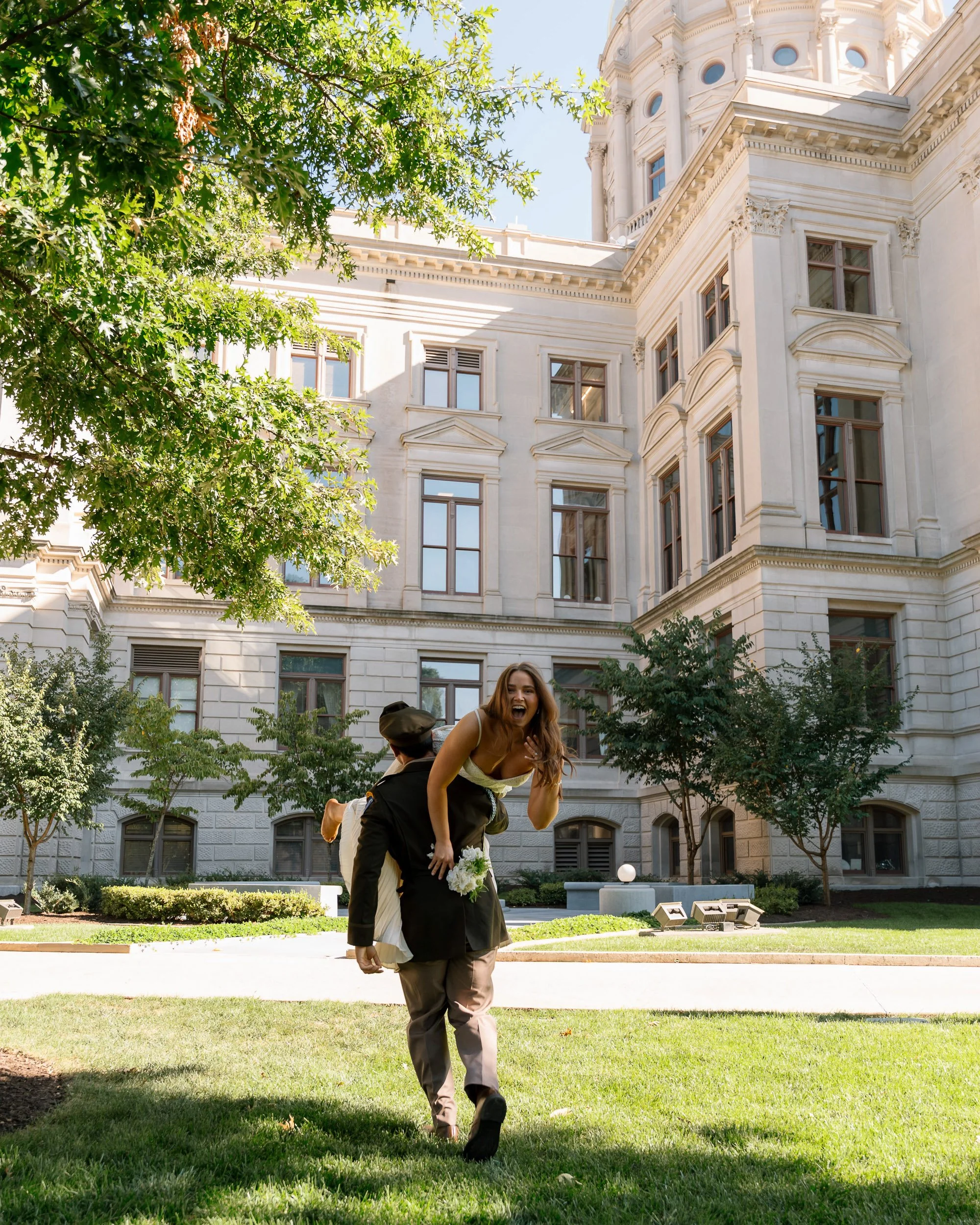 A joyful couple in formal attire, with the woman laughing and the man carrying her on his back, outdoors in front of a historic building with large windows, trees, and a lawn.