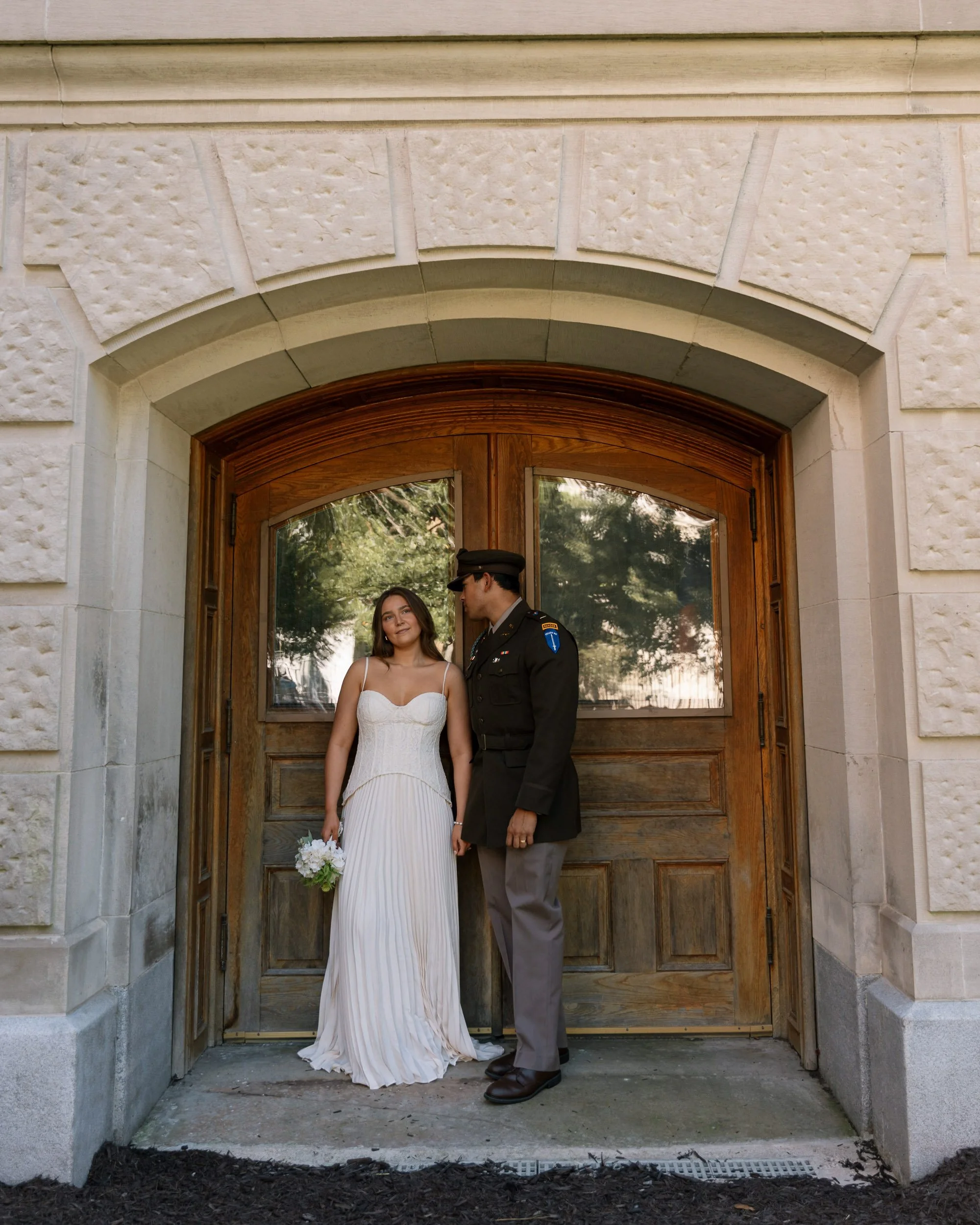 A bride in a white dress holding a bouquet standing next to a man in a formal military uniform in front of a wooden door.