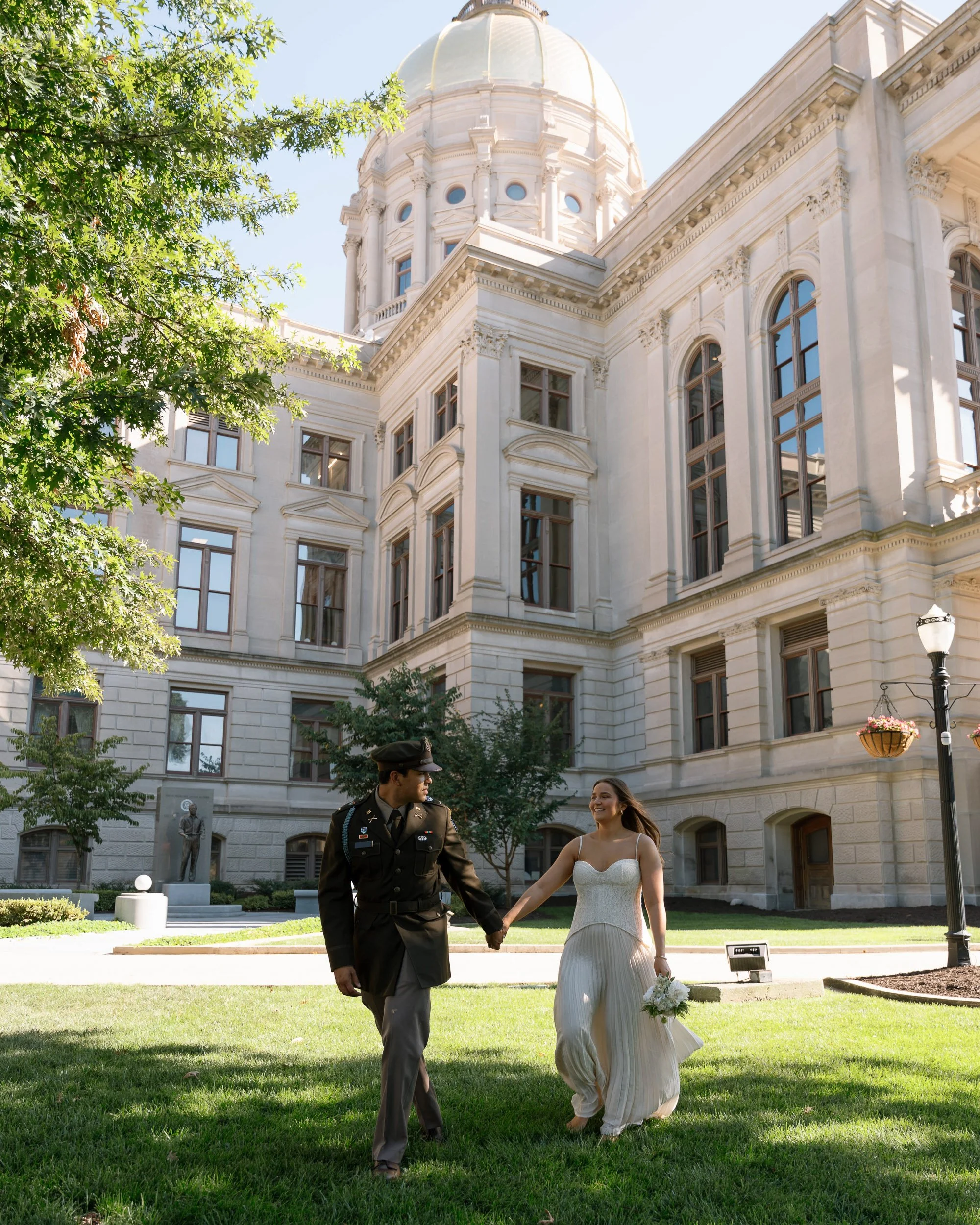 A woman in a white wedding dress holding a bouquet and a man in a military uniform holding hands walking on a lawn outside a large historic white building with a domed roof and tall windows.