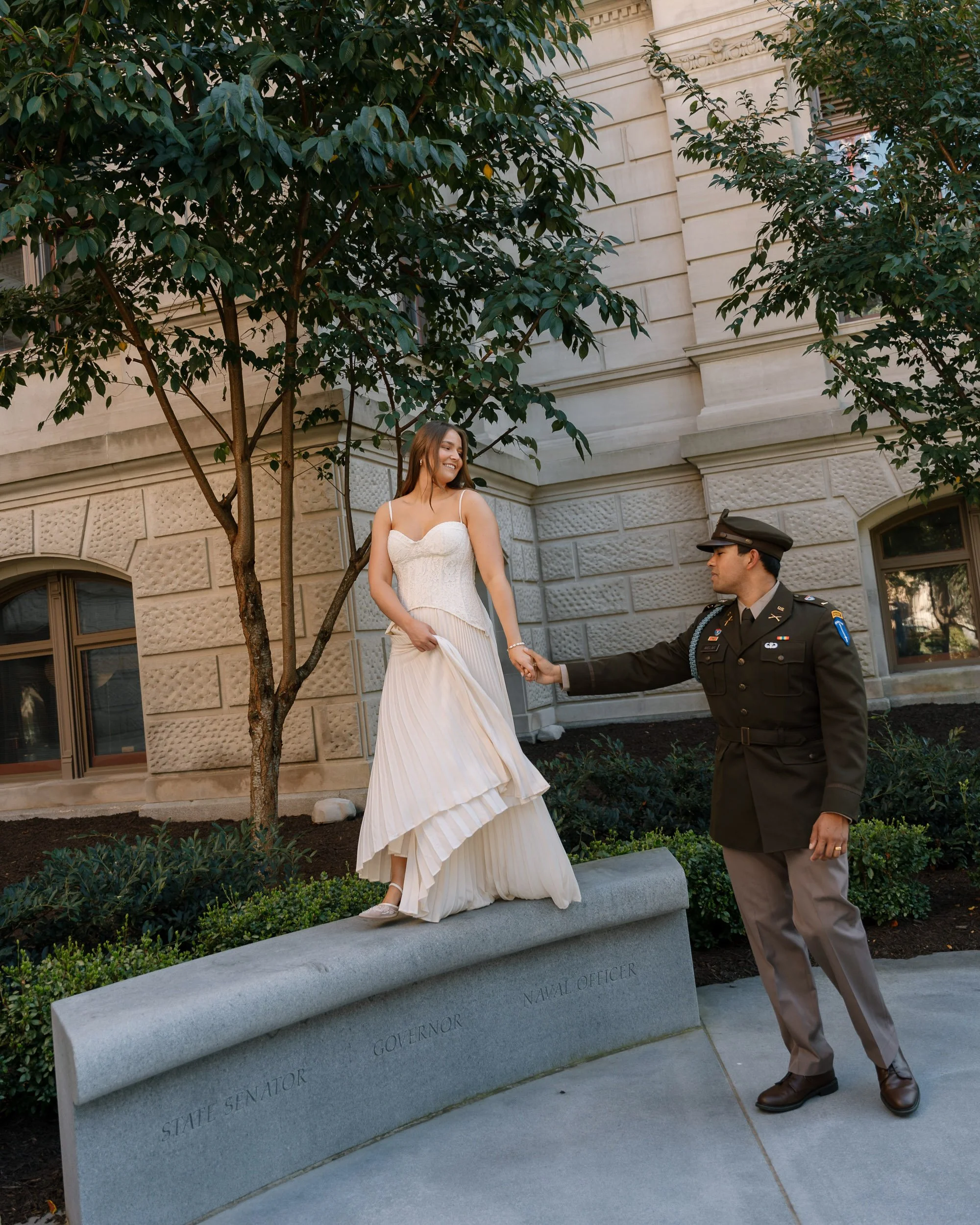 A woman in a white dress standing on a stone bench labeled "State Senator," "Governor," and "Navy Officer," holding hands with a uniformed navy officer in a formal uniform, outdoors near a stone building and trees.