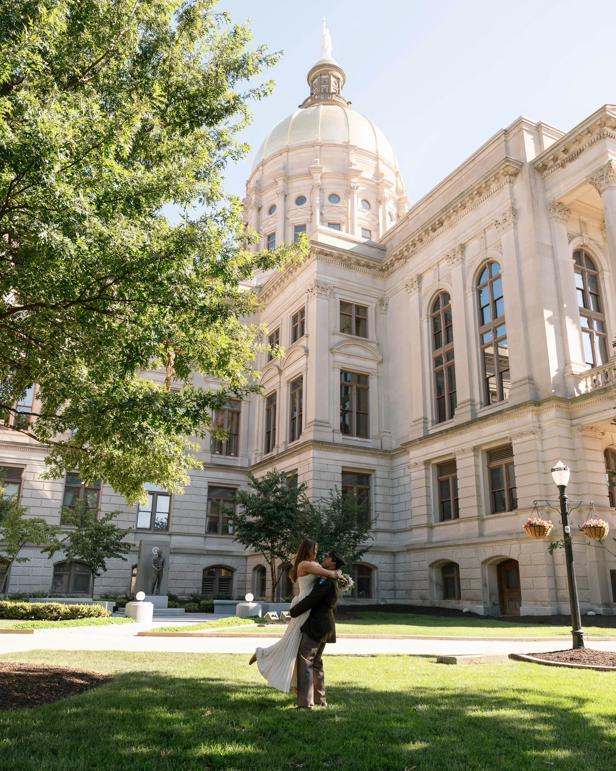 A couple dancing on a grassy area in front of a historic white building with a large dome, surrounded by trees and a lamppost with hanging flower baskets.