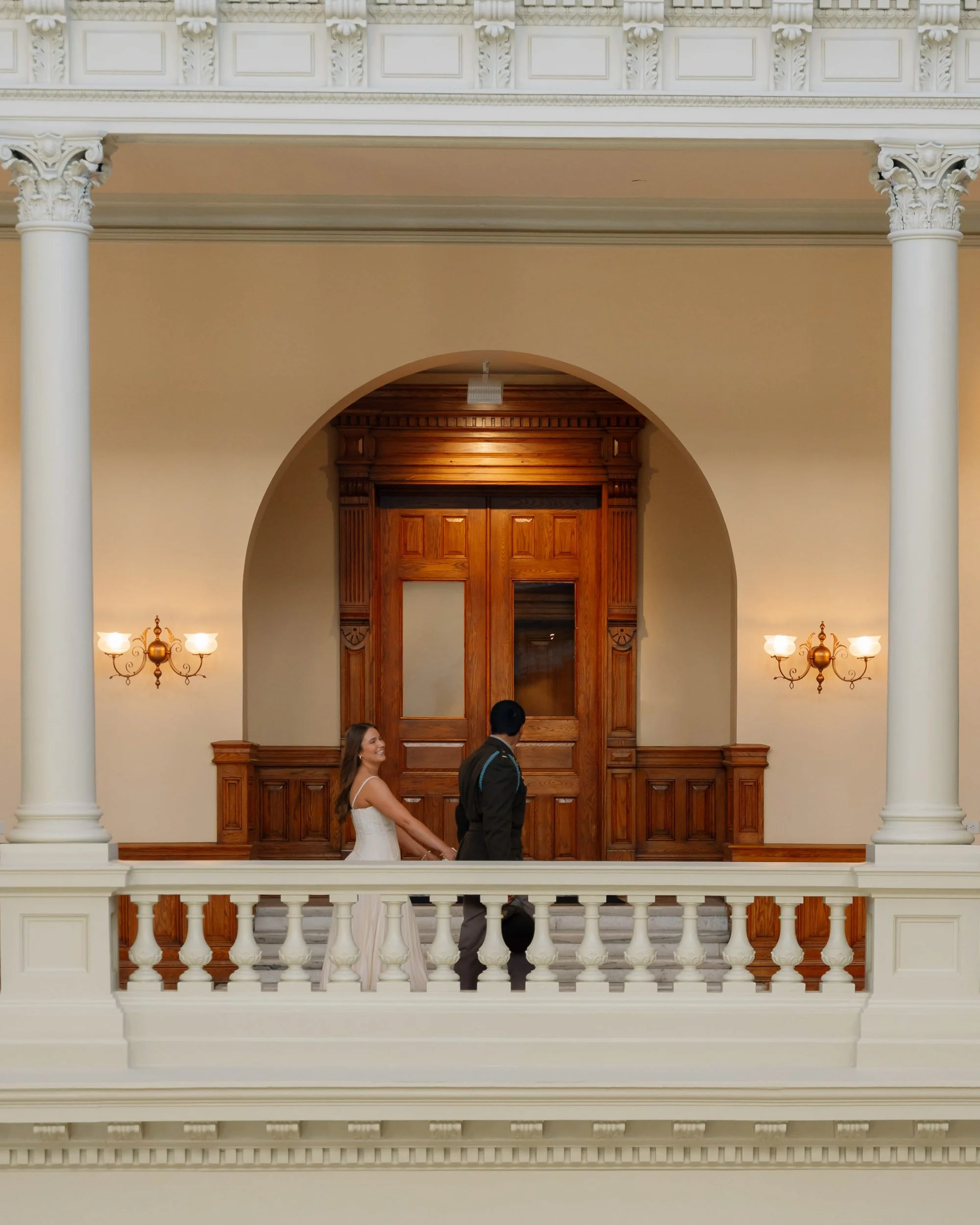 A bride and groom holding hands on a balcony inside a grand, historic building with ornate columns, wooden paneling, and decorative lighting.