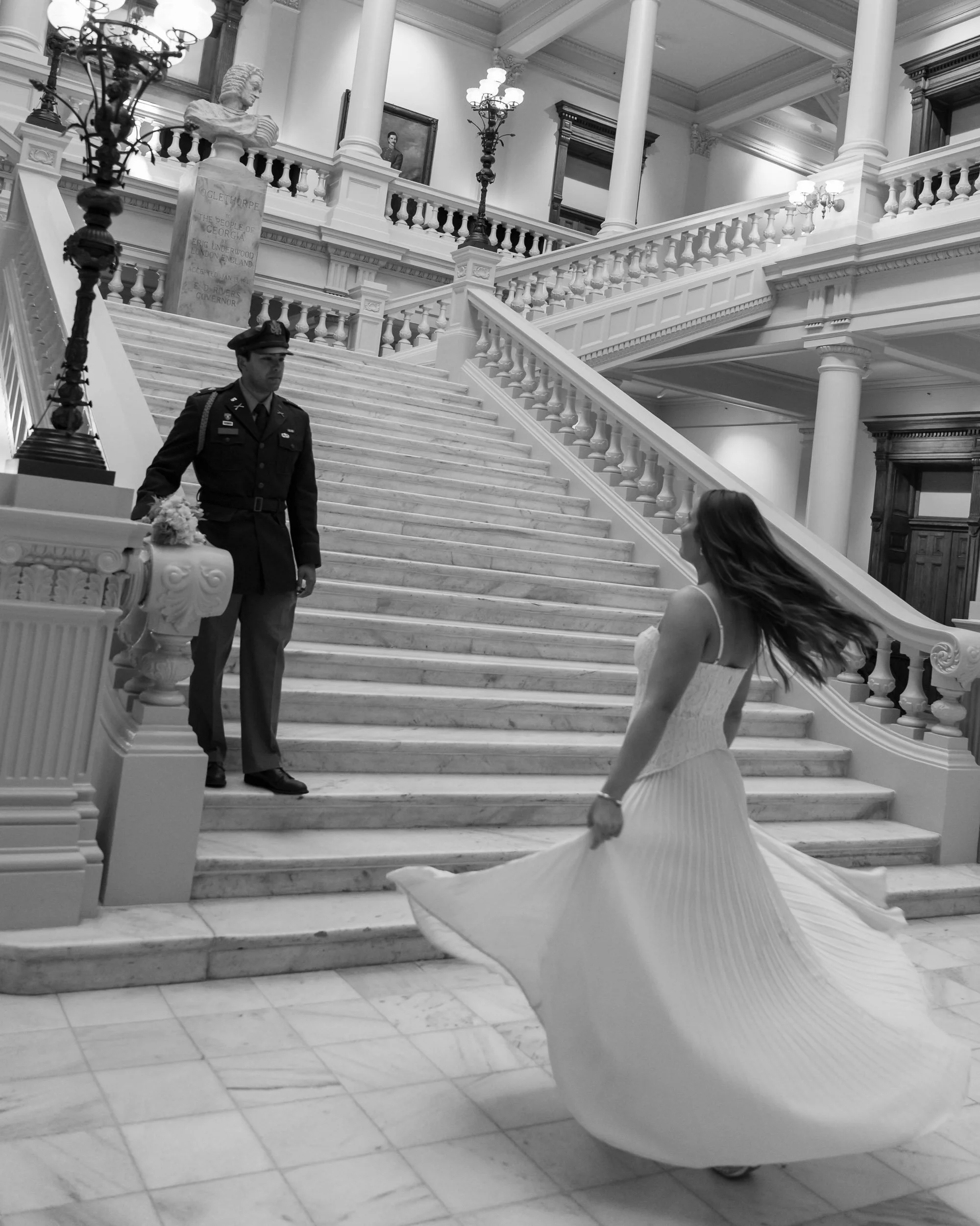 A woman in a flowing dress stands at the bottom of a grand staircase, with a police officer in uniform standing on the stairs above her. The staircase is ornate, with marble steps and decorative railings, inside a formal building with tall columns an