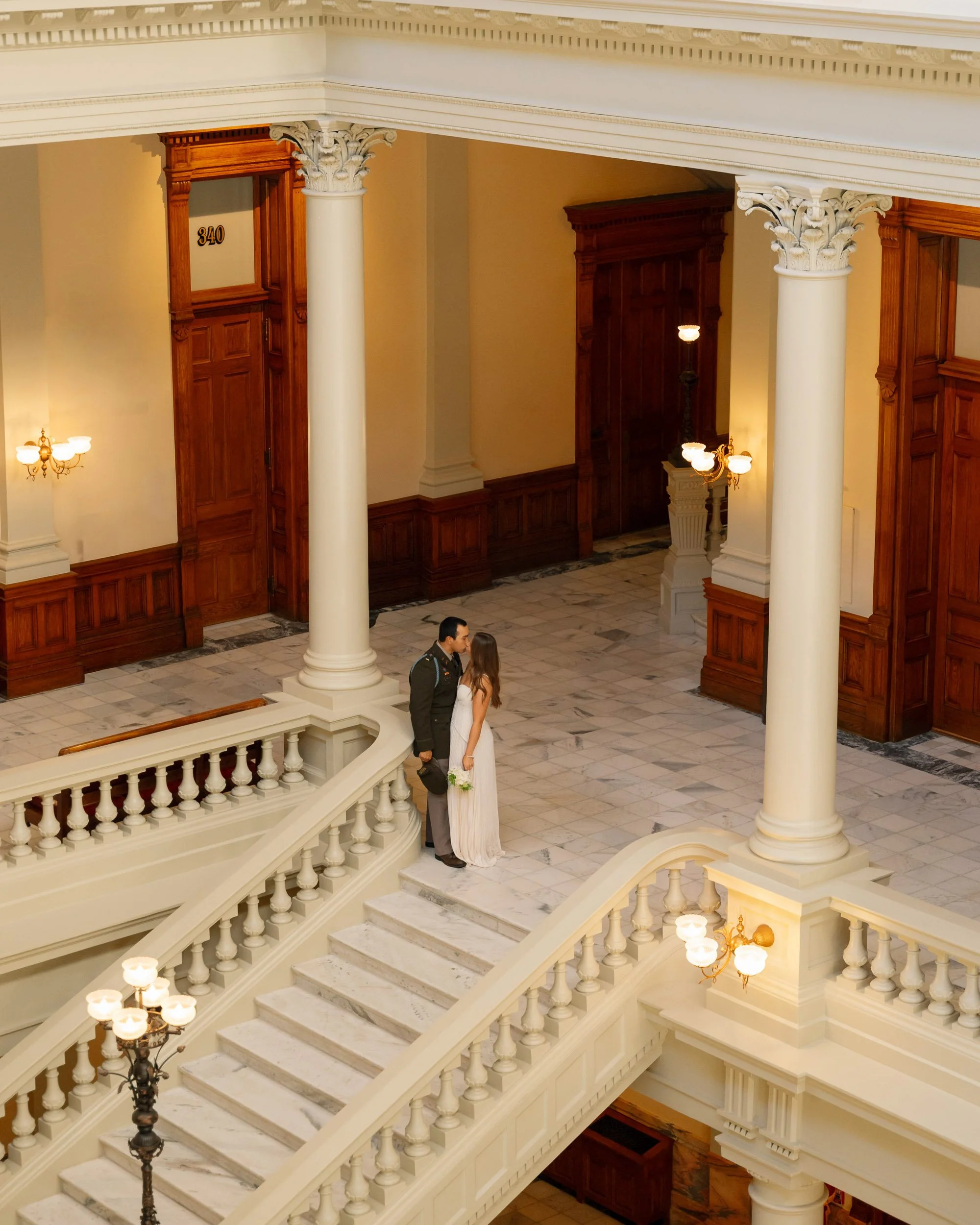 A man in formal attire and a woman in a white dress stand close together on a staircase inside a grand building with wood paneling, marble floors, and ornate columns, sharing an intimate moment.