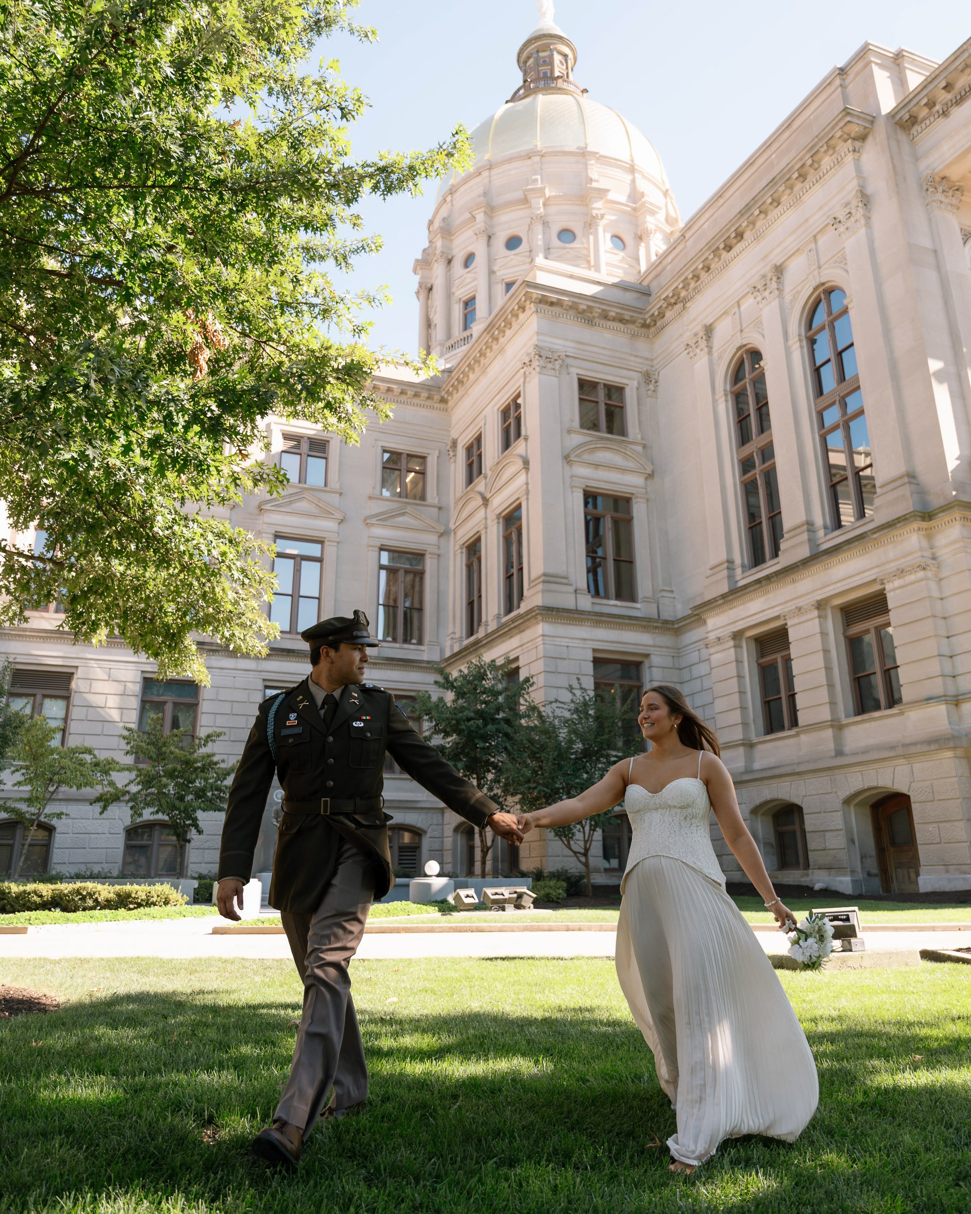 A couple, a woman in a white dress holding a bouquet, and a man in a military uniform, holding hands and walking in front of a large historic building with a dome, trees, and a sunny sky.
